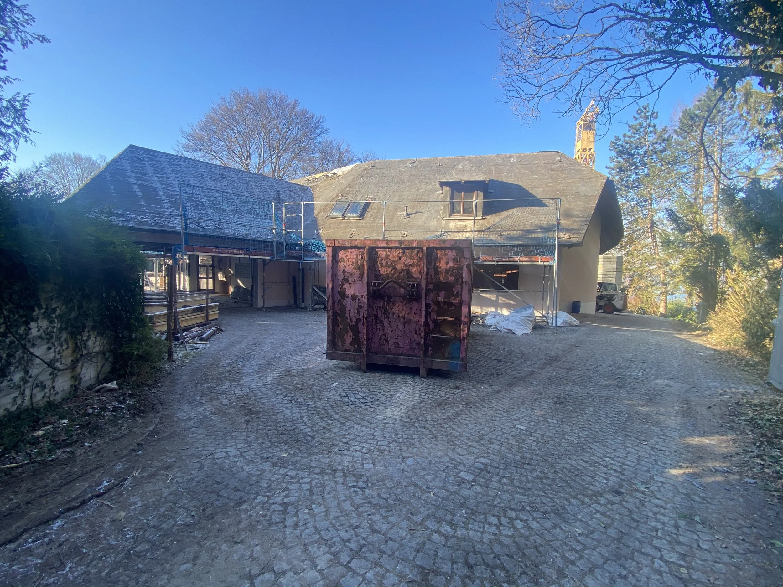Baustelle vor einem Haus mit Gerüst, im Vordergrund eine alte, kaputte Mülltonne, Hintergrund Bäume und blauer Himmel