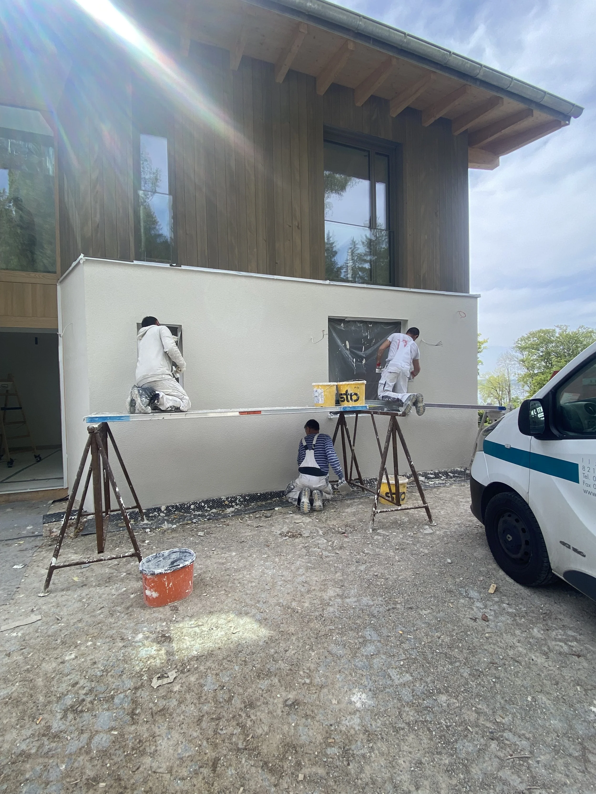 Drei Bauarbeiter streichen die Außenwand eines Gebäudes. Zwei sind auf Gerüsten, während der dritte auf den Boden kniet. Es gibt Werkzeuge, Eimer und einen weißen Dienstwagen neben der Baustelle.