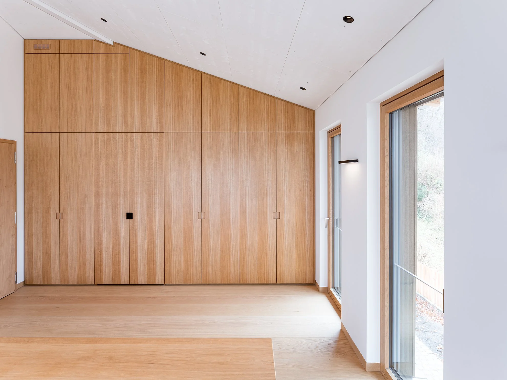 Innenaufnahme eines modernen Raums mit Holzvertäfelung an der Wand, weißen Wänden, Holzboden, einem kleinen schwarzen Wandlicht und großen Fenstern mit Holzbalken, die Tageslicht in den Raum lassen.