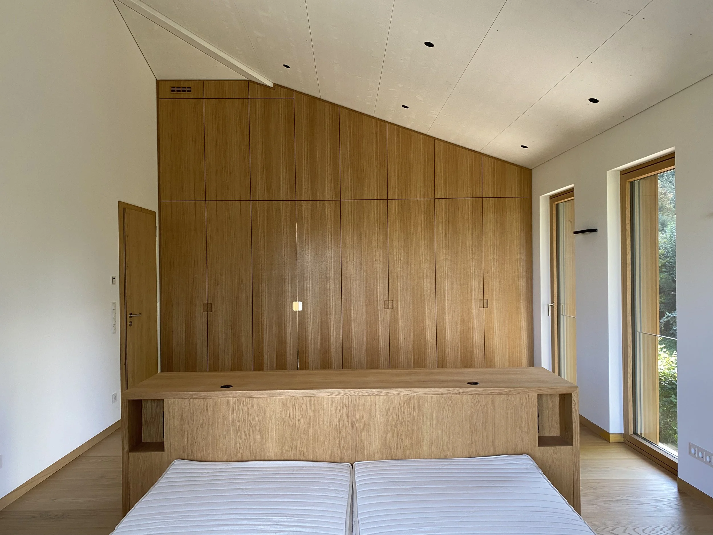 Fotografie eines minimalistisches Schlafzimmers mit weißen Wänden, Holzboden, großen Fenstern mit Blick auf Bäume, Holzmöbeln und einer Holzwand. Kein Bett, sondern zwei zusammengeschobene Matratzen.