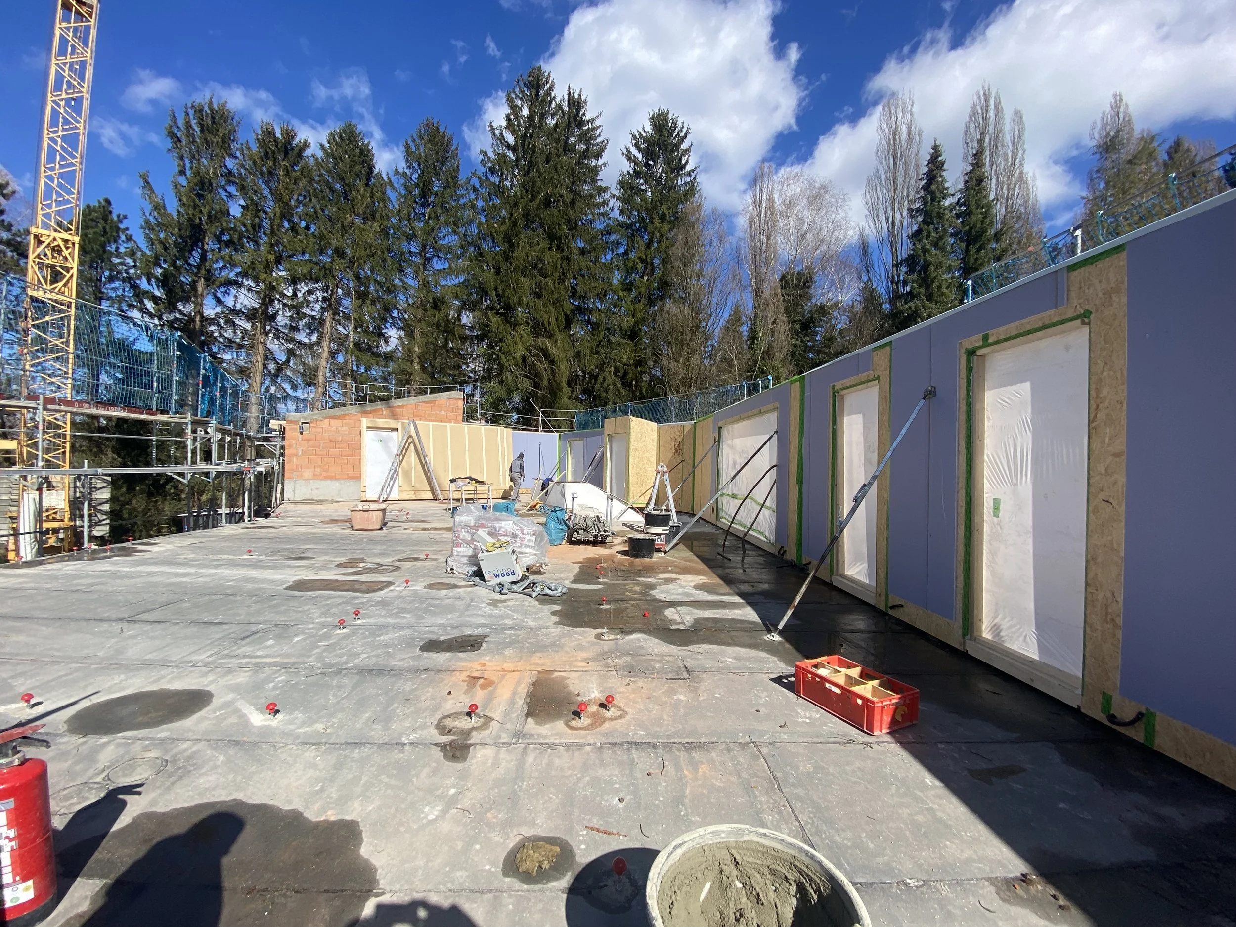 Bauarbeiter arbeiten an einer Baustelle im Freien, umgeben von Baugerüsten, Bau-materialien und Werkzeugen, während sich im Hintergrund ein Wald mit hohen Bäumen befindet.