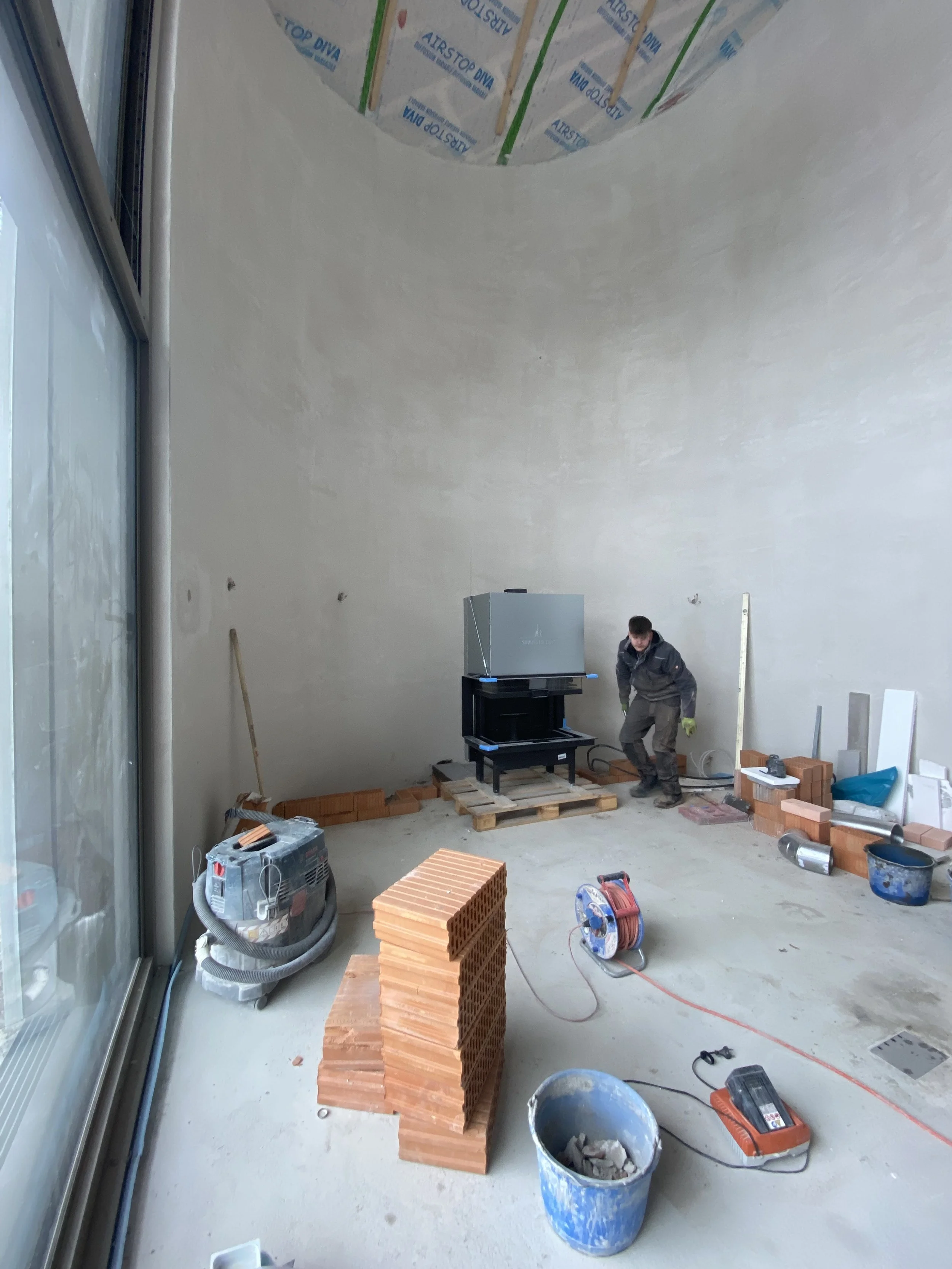 Innenaufnahme eines Raumes im Bau, ein Arbeiter steht auf einer noch unvollendeten Bodenfläche, umgeben von Baumaterialien wie Ziegelsteinen, Werkzeugen und Kabeln, mit einer großen Fensterscheibe auf der linken Seite.