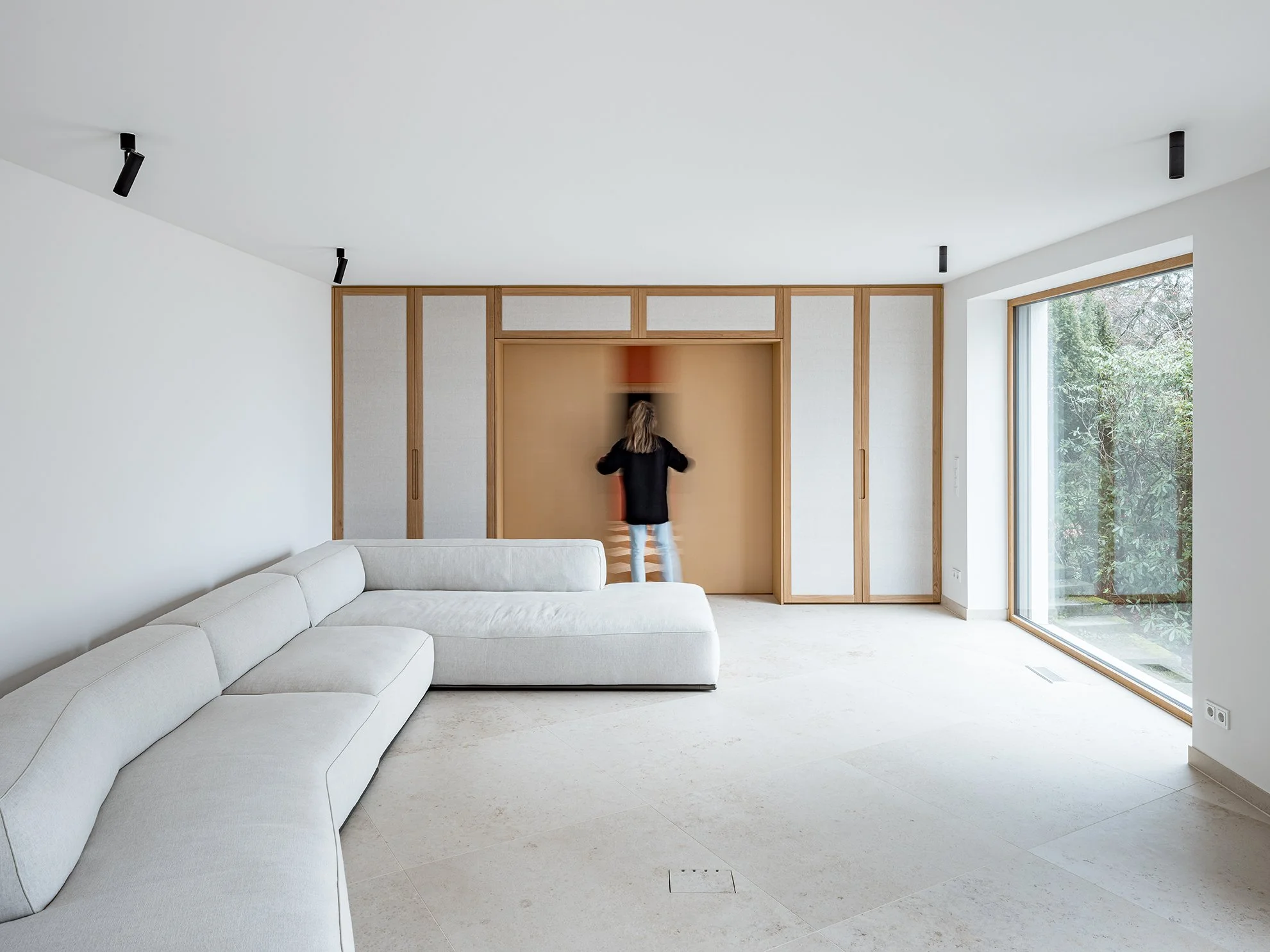 Person öffnet einen Holzschrank in einem modernen, hellen Wohnzimmer mit großen Fenstern, weißen Wänden und einem weißen Sofa.