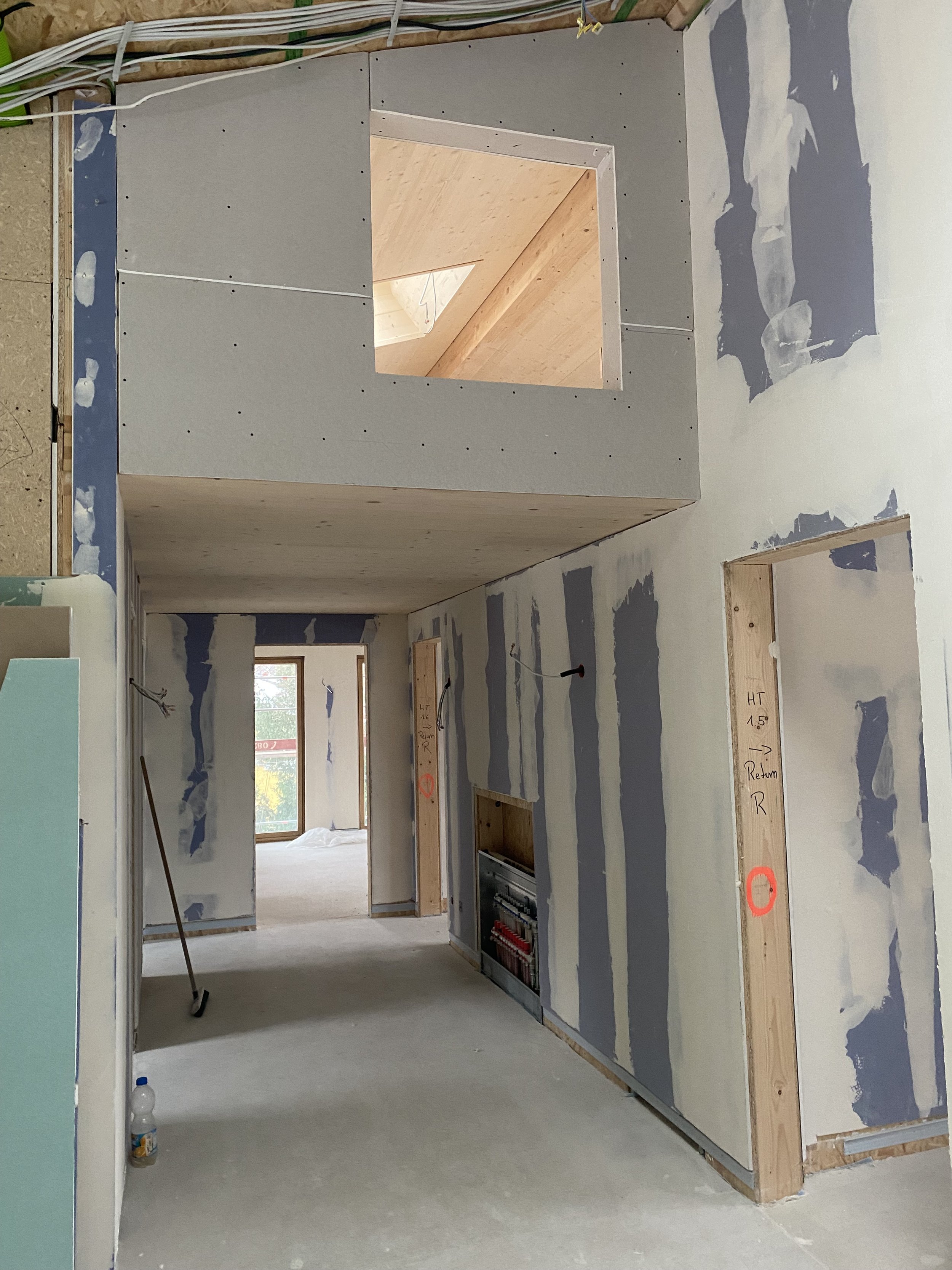 Innenansicht eines Hauses im Bau, mit unvollständiger Trockenbauwand, offene Fenster, Bauwerkzeuge und Rohre sichtbar.