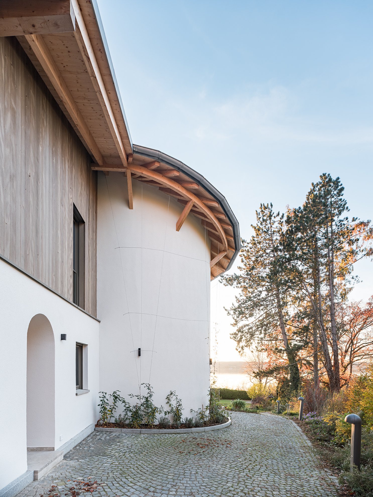 Modernes Haus mit weißer Fassade, Holzverkleidung und Rundbogen-Tür, umgeben von einem gepflasterten Weg und Bäumen im Herbst, Sonnenuntergang im Hintergrund.
