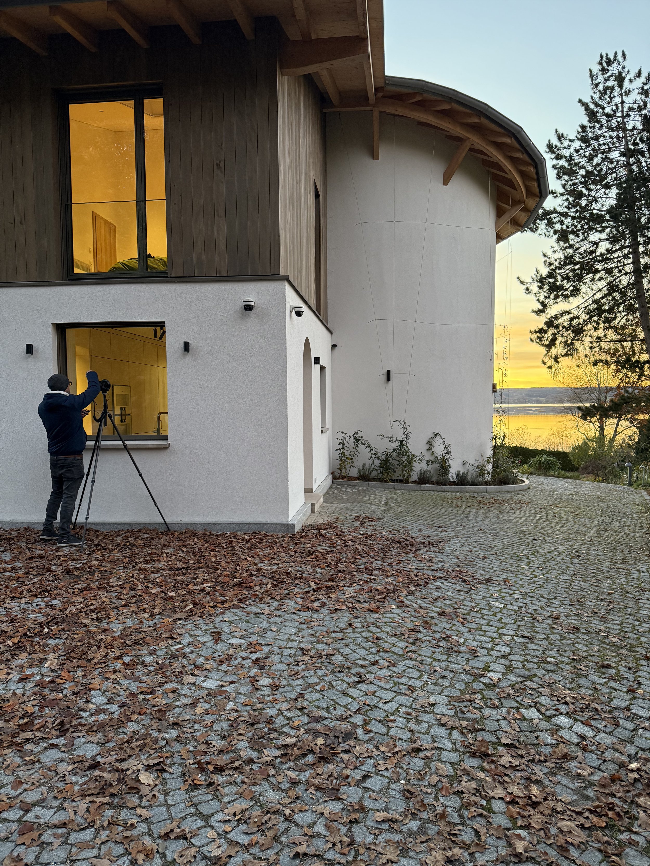 Ein Mann macht Foto mit einer Kamera auf einem Stativ vor einem modernen Haus bei Sonnenuntergang. Das Haus hat weiße und holzvertäfelte Außenwände, große Fenster und ist von einem gepflasterten Weg umgeben. Im Hintergrund ist ein Baum und ein Gewäss