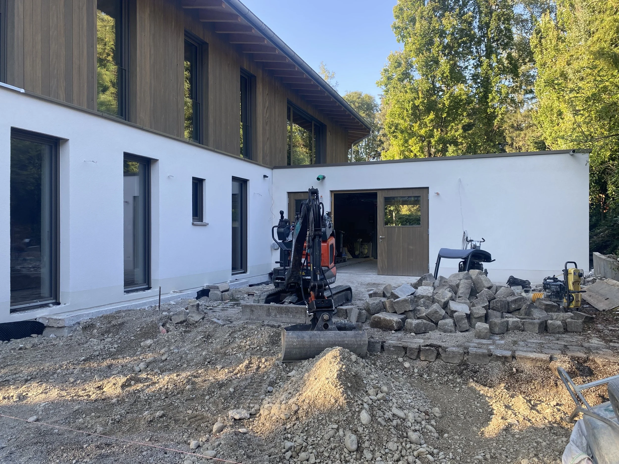 Baustelle vor einem Mehrfamilienhaus mit weißen und holzverkleideten Fassaden, im Vordergrund befinden sich Baustoffe, Steine und Baugeräte.