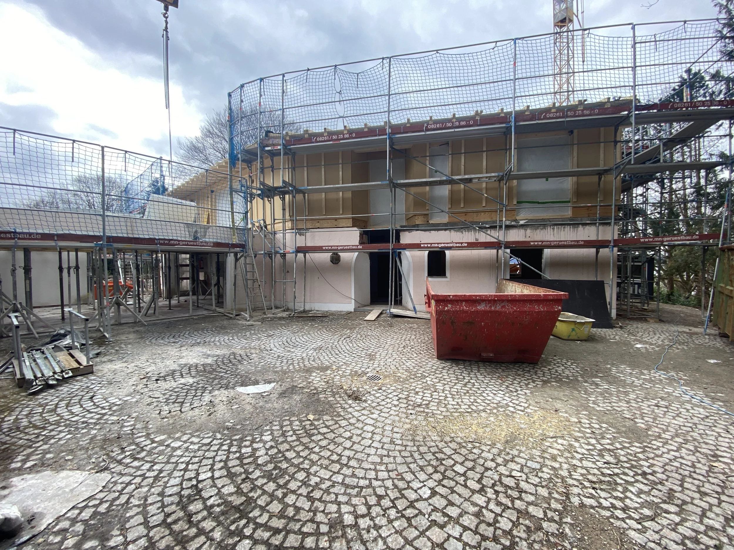 Baustelle eines Hauses mit Gerüsten und Baugeräten, im Vordergrund ein roter Container und eine gelbe Schubkarre, die Baustelle befindet sich auf einer gepflasterten Fläche, im Hintergrund sind Bäume und ein Himmel mit Wolken.