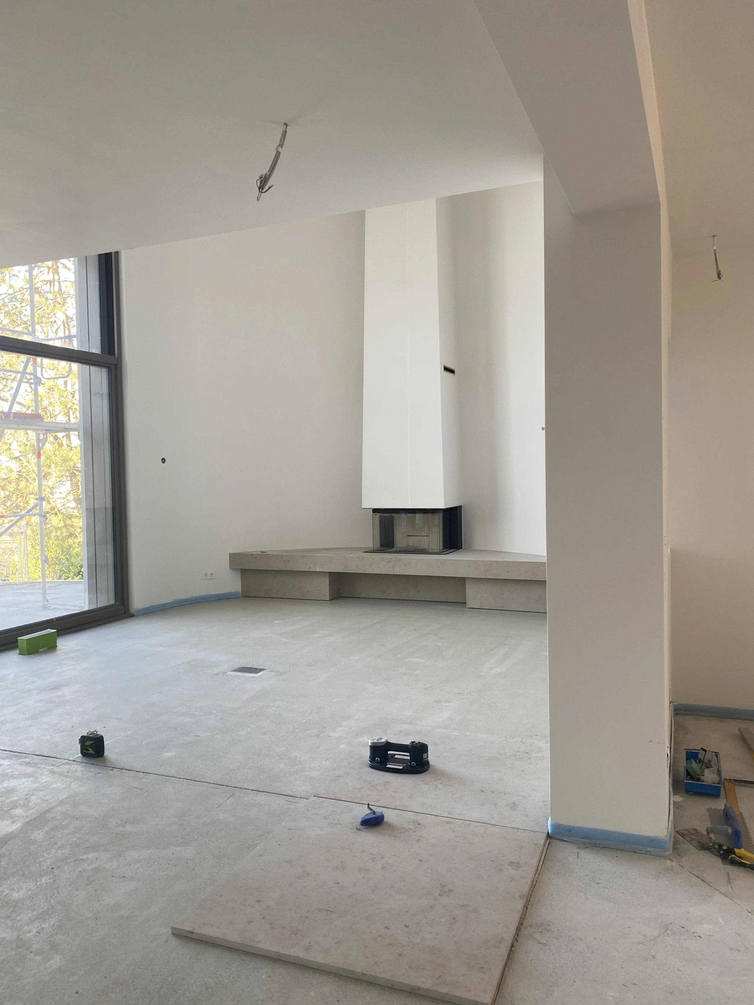 Innenraum eines noch unvollständigen modernen Hauses mit offener Raumgestaltung, Kamin, große Fensterfront und Baustellengeräte auf dem Boden.