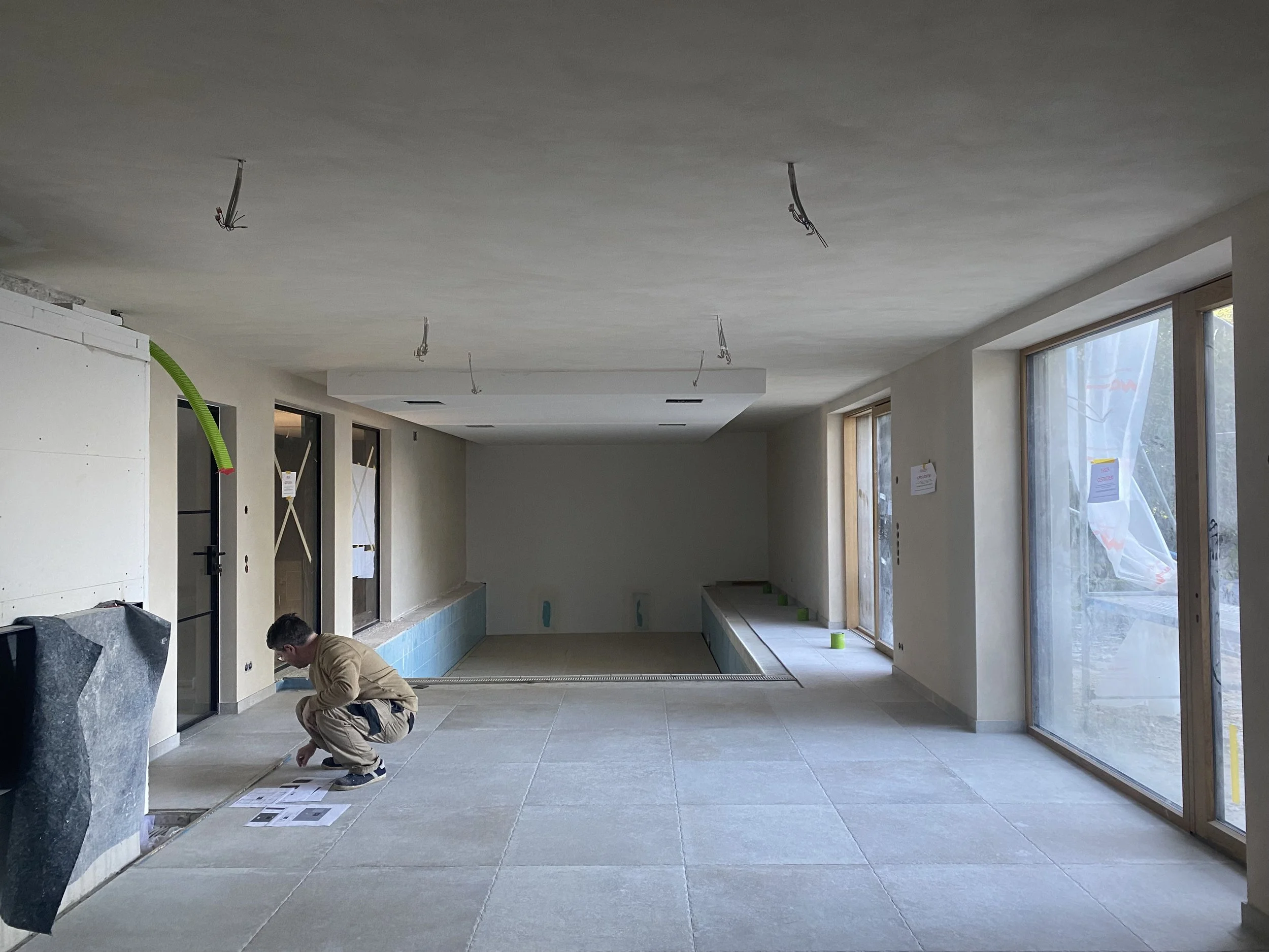 Innenraum eines noch incomplete Hauses mit einem Mann, der auf dem Boden arbeitet, große Fenster und unvollendete Wände.