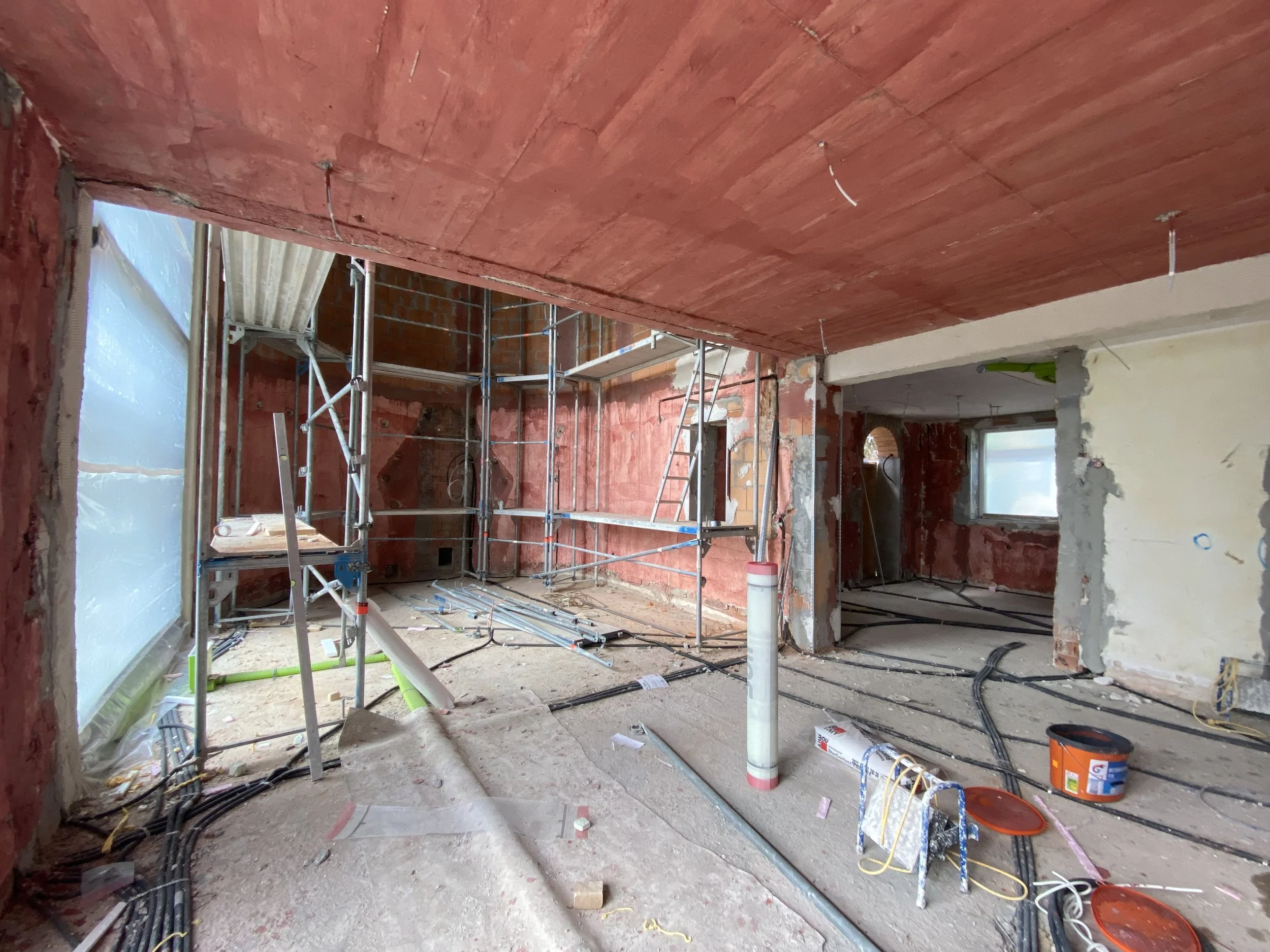 Innenraum eines im Bau befindlichen Hauses mit Gerüsten, Rohren und Bauwerkzeugen, Wänden in roter und weißer Farbe, Baustoffen und Kabeln auf dem Boden.