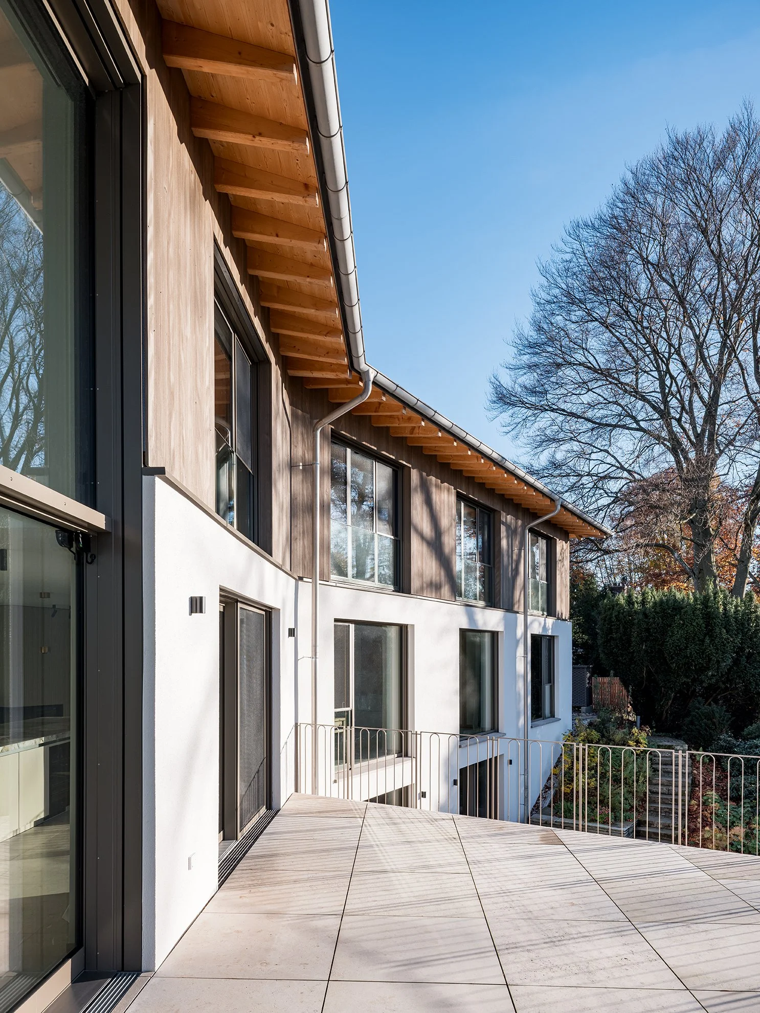 Moderner Wohnhaus mit Balkon, große Fenster, Holz- und Putzfassade, Baum im Hintergrund, blauer Himmel, Sonnenlicht, Außenbereich