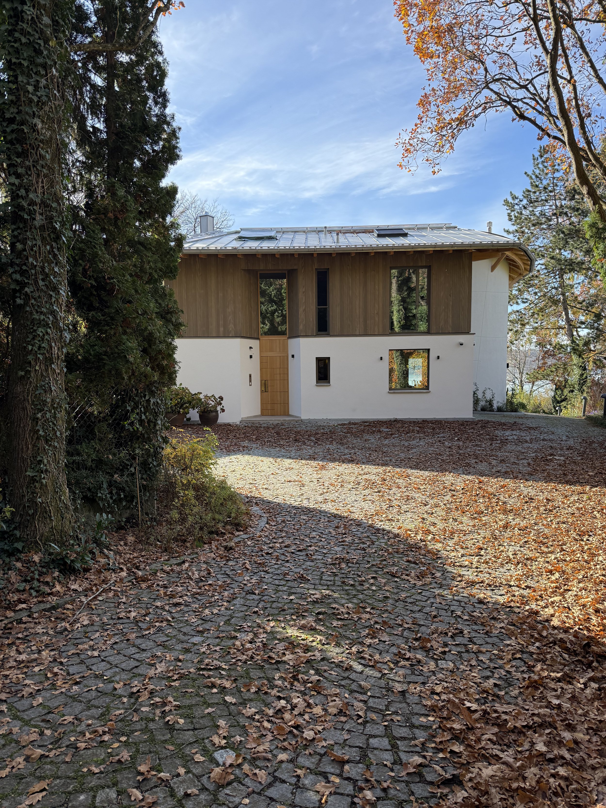 Moderne zweistöckige Villa mit Holz- und Weißfassade, große Fenster, Schindeldach und Kiesweg im herbstlichen Garten mit Laub.