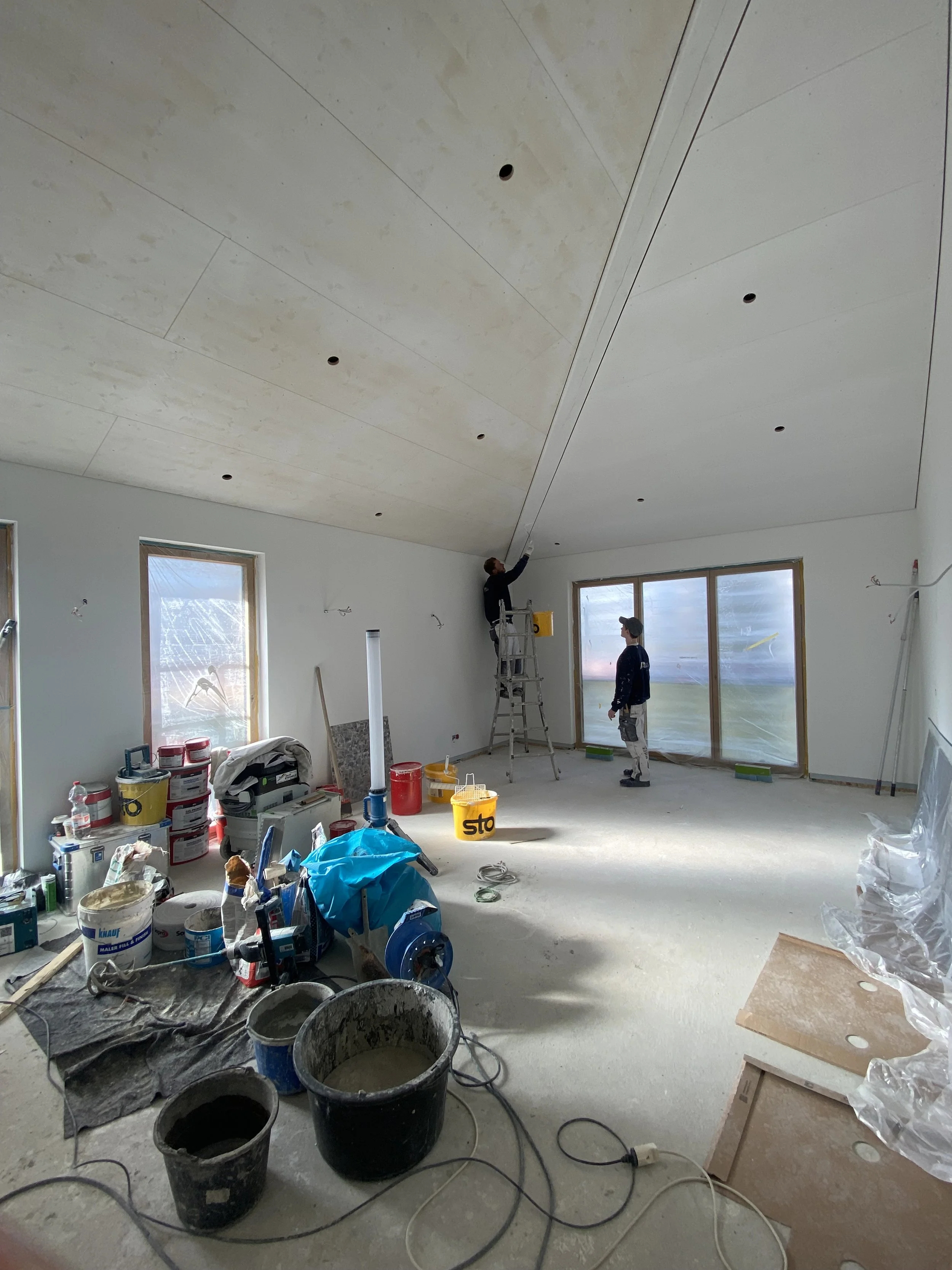 Zwei Arbeiter streichen die Decke eines Raumes während einer Renovierung, umgeben von Baumaterialien und Werkzeugen.