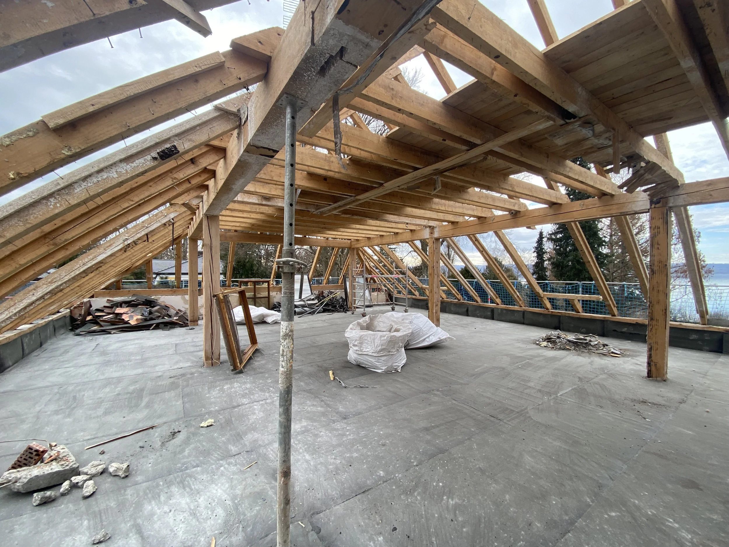 Bauarbeiten auf einem Dach mit Holzkonstruktion und Stahlstützen, sichtbarer Dachbodenboden, im Hintergrund Bäume und Himmel.