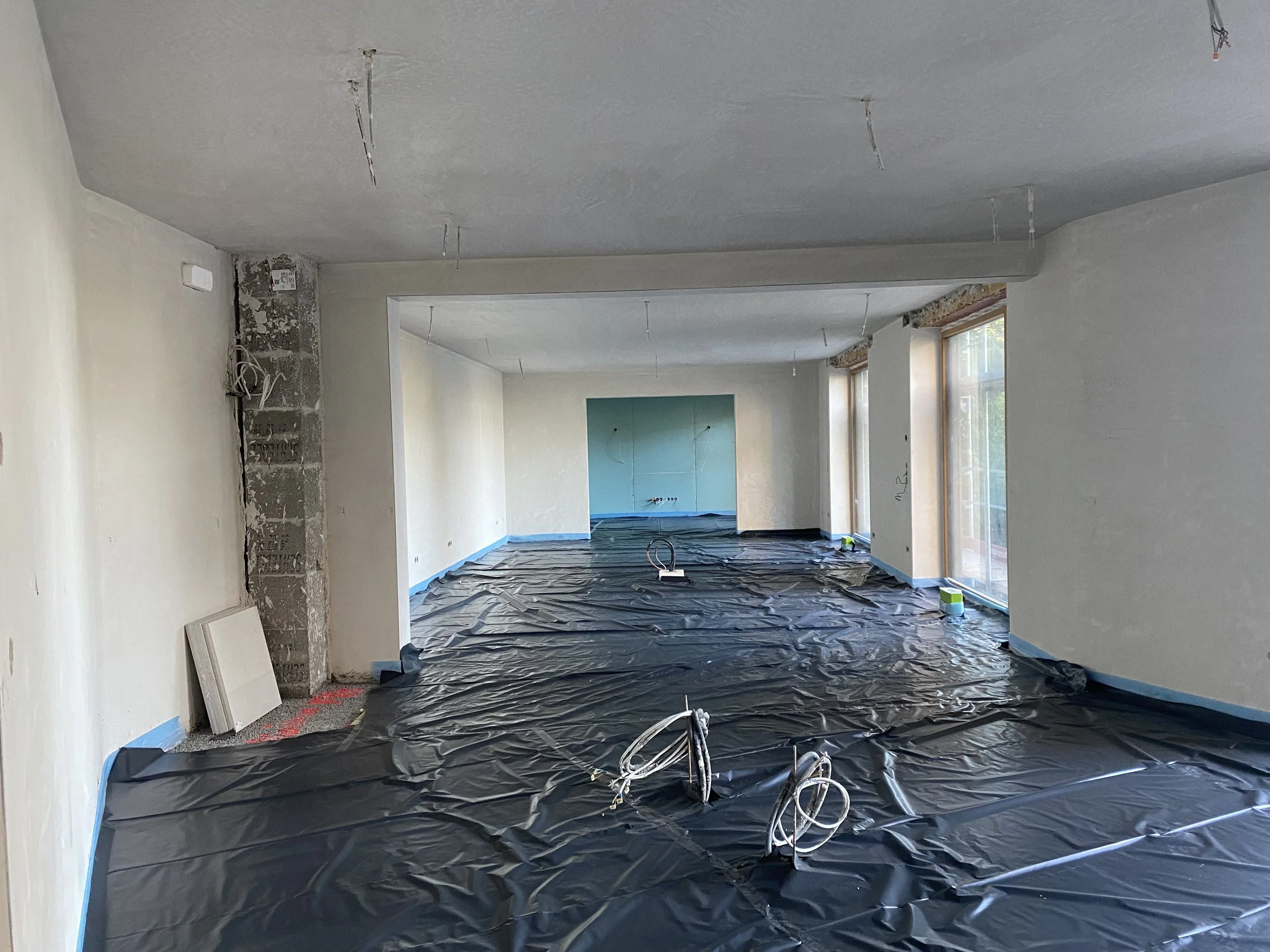 Innenraum eines Renovierungsprojekts mit weiß gestrichenen Wänden, große Fenster, schwarzer Schutzfolie auf dem Boden, Kabel und Rohre hängen von der Decke.