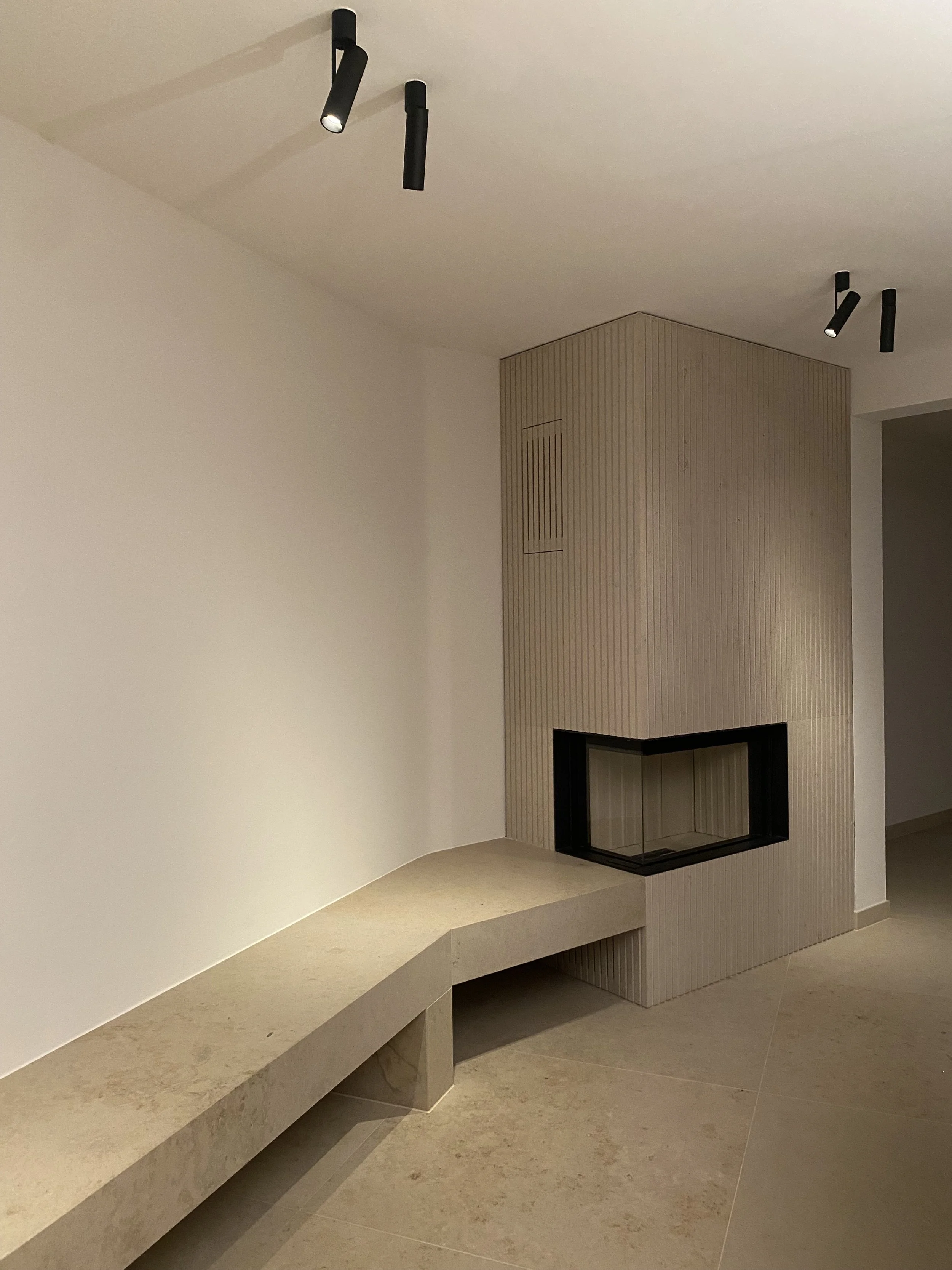 Moderner Kamin mit beige-gemusterter Verkleidung und einer gebogenen Sitzbank aus hellem Stein oder Beton darunter, in einem minimalistischen Raum mit weißen Wänden und schwarzen Deckenleuchten.