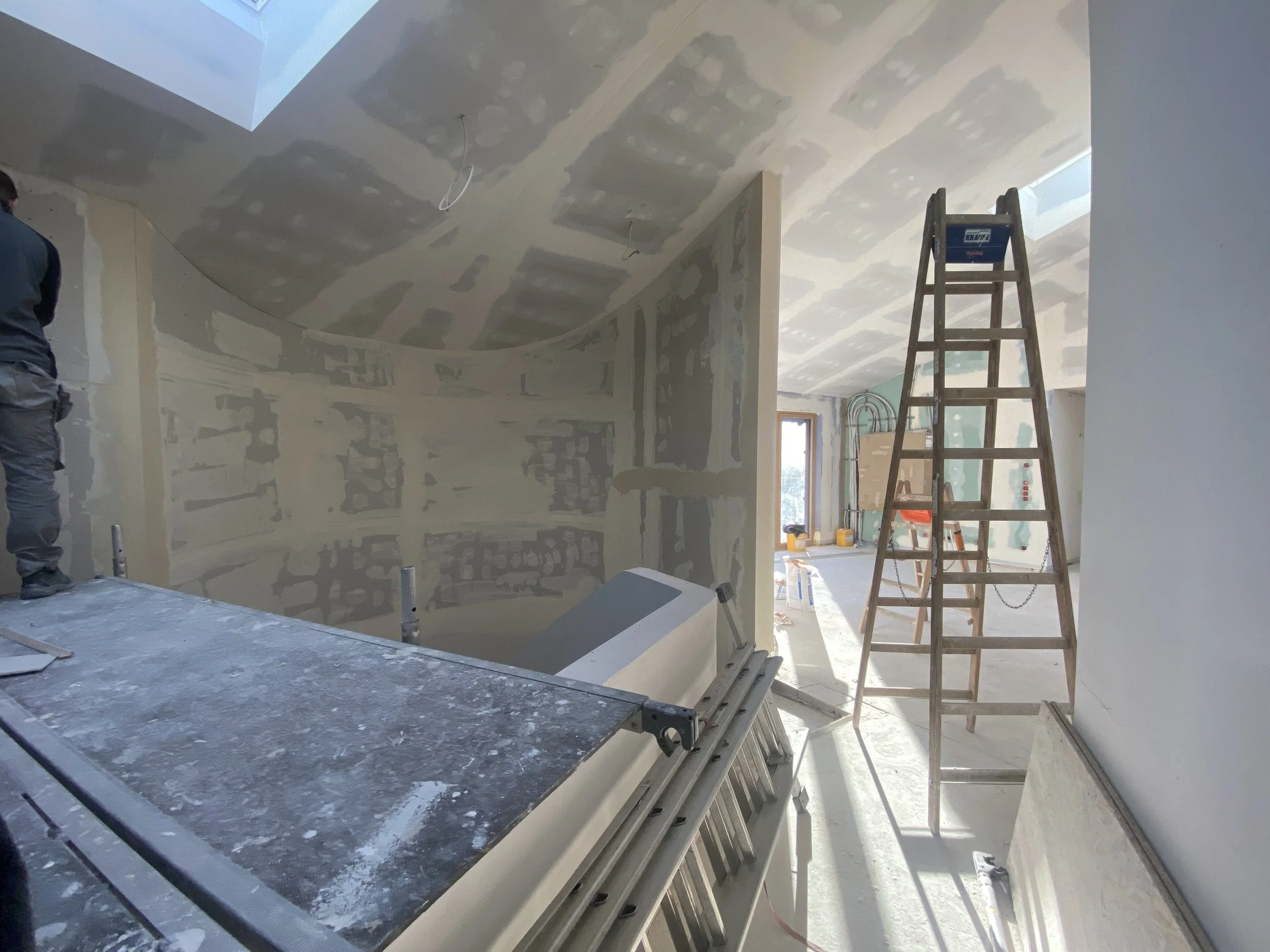 Innenraum eines im Bau befindlichen Hauses mit geöffnetem Dachfenster, unvollendeten Wänden und einer Leiter, die in einem Raum mit vielen Baumaterialien steht.