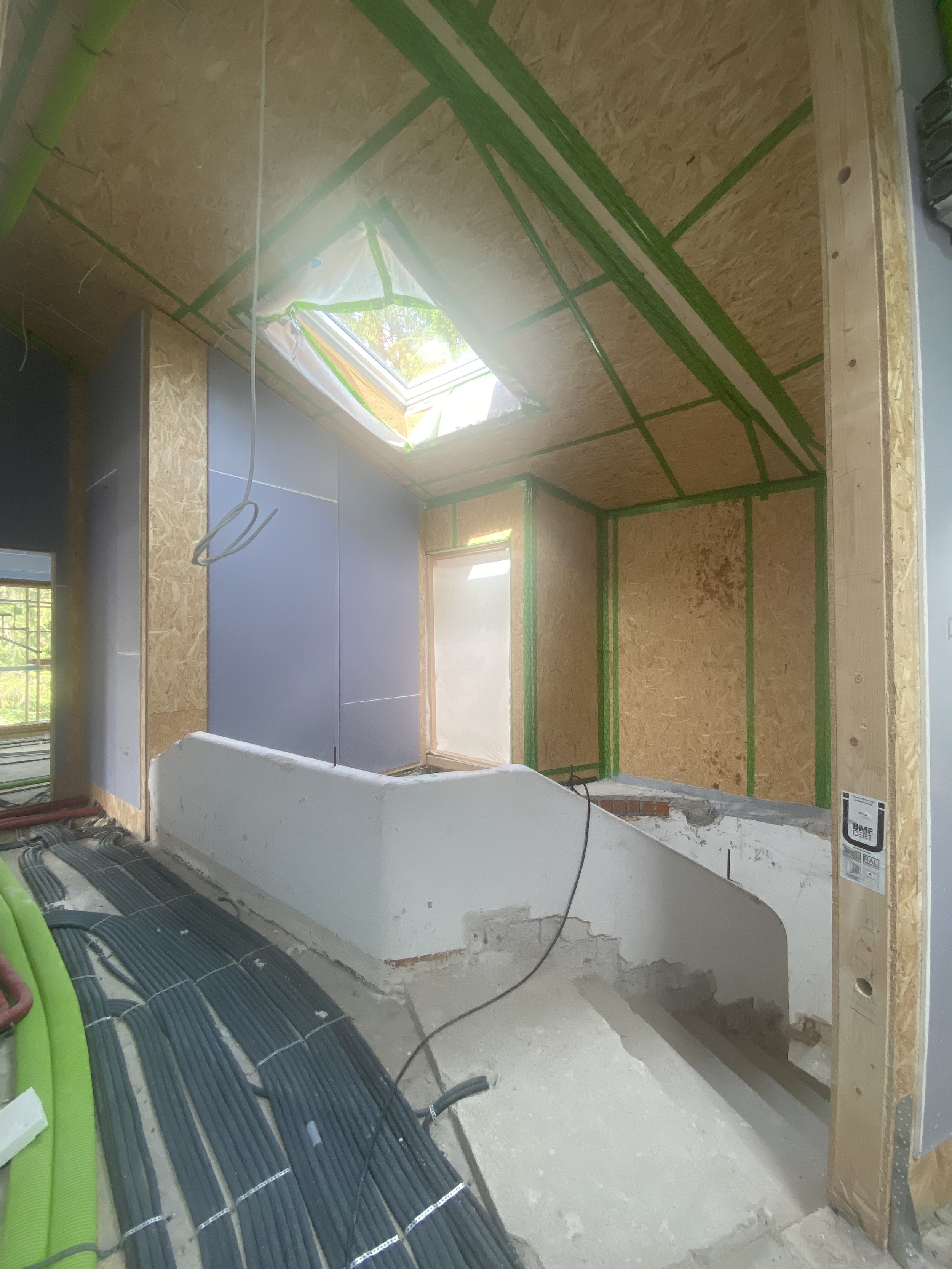 Innenraum eines Hauses während der Bauarbeiten mit rohem Holz und Isolierung, Kabeln und Rohren auf dem Boden, und einem Dachfenster, das Tageslicht hereinlässt.