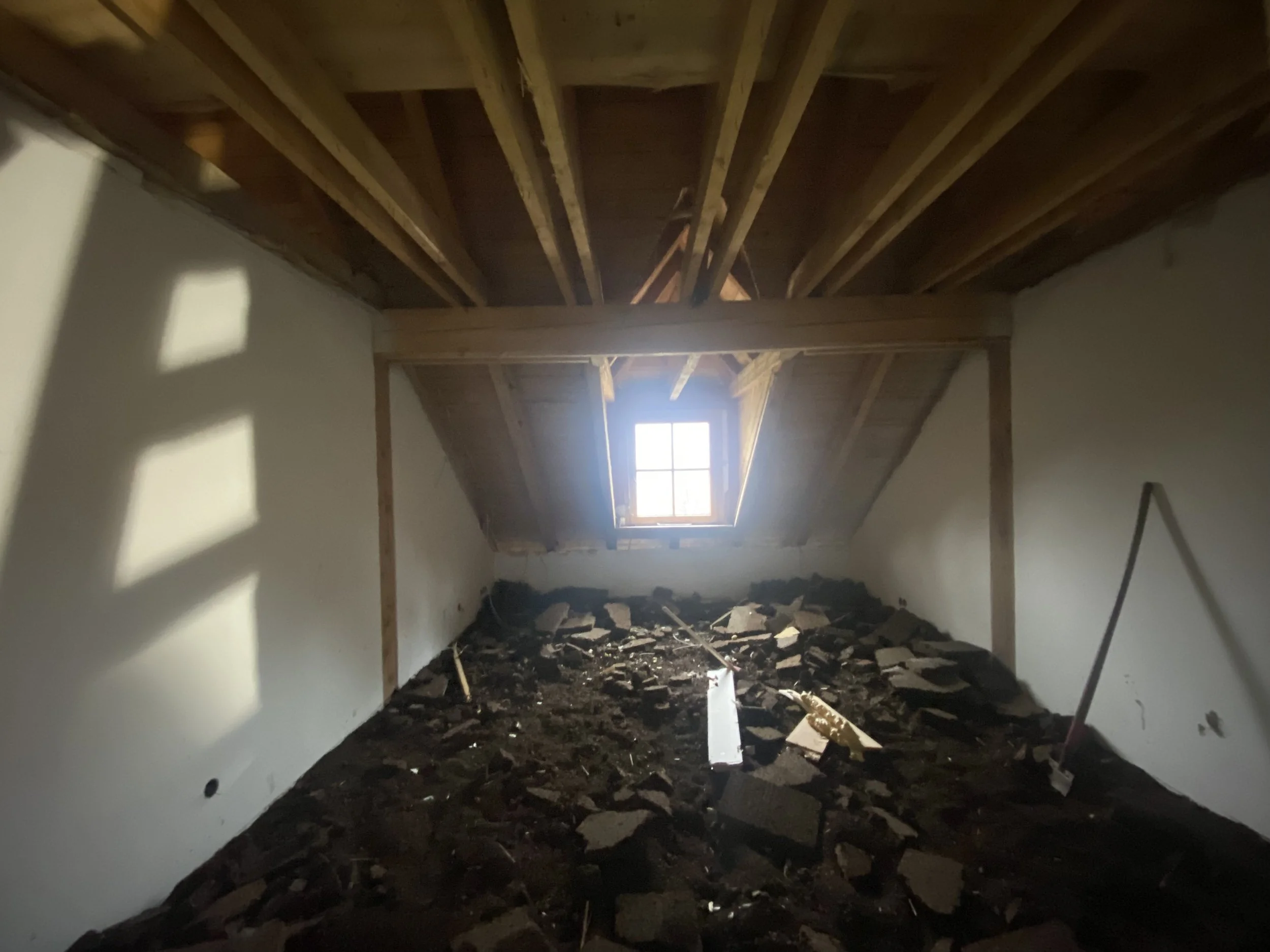 Ein unvollendeter Dachraum mit Holzbalken, einer kleinen Fensteröffnung und Baumaterial auf dem Boden.