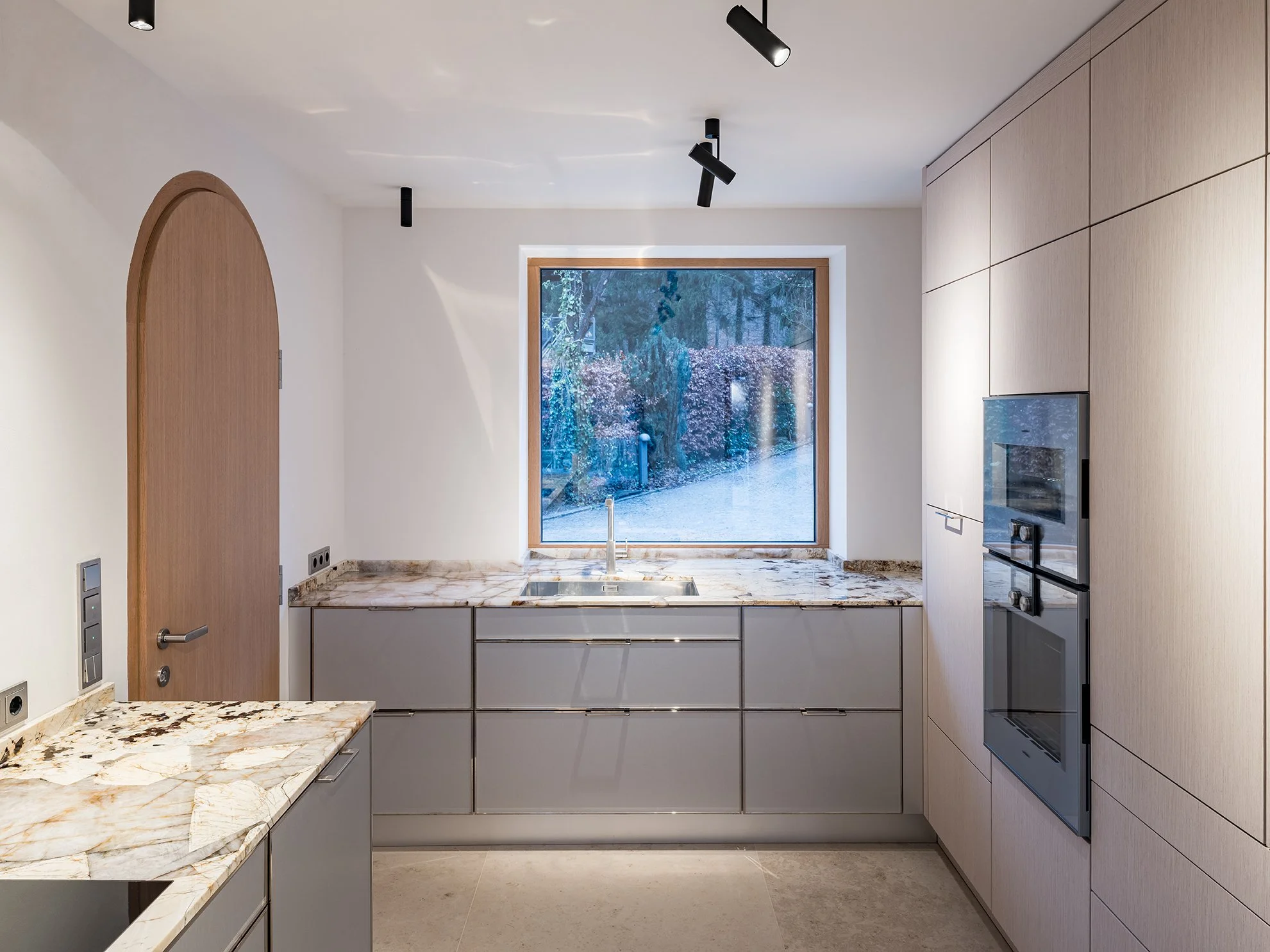 Moderne Küchenschrankküche mit großen Keramikplatten, Blick auf einen Garten mit Bäumen und Hecke durch das Fenster.