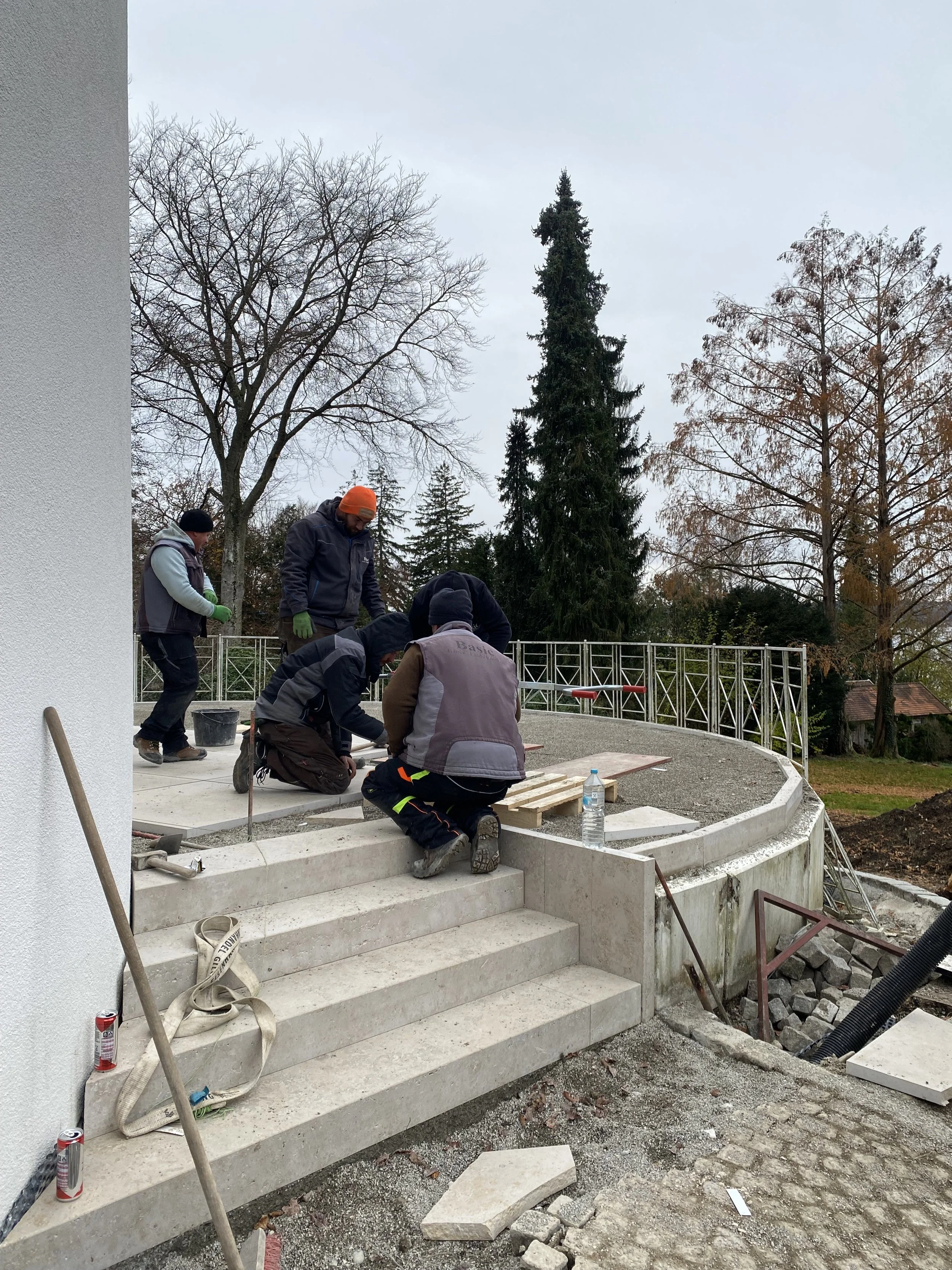 Menschen arbeiten an einer Baukrippe mit Treppe im Freien, einige bücken sich und montieren Steine, bei kühlem Wetter mit bewölktem Himmel, Bäumen im Hintergrund.