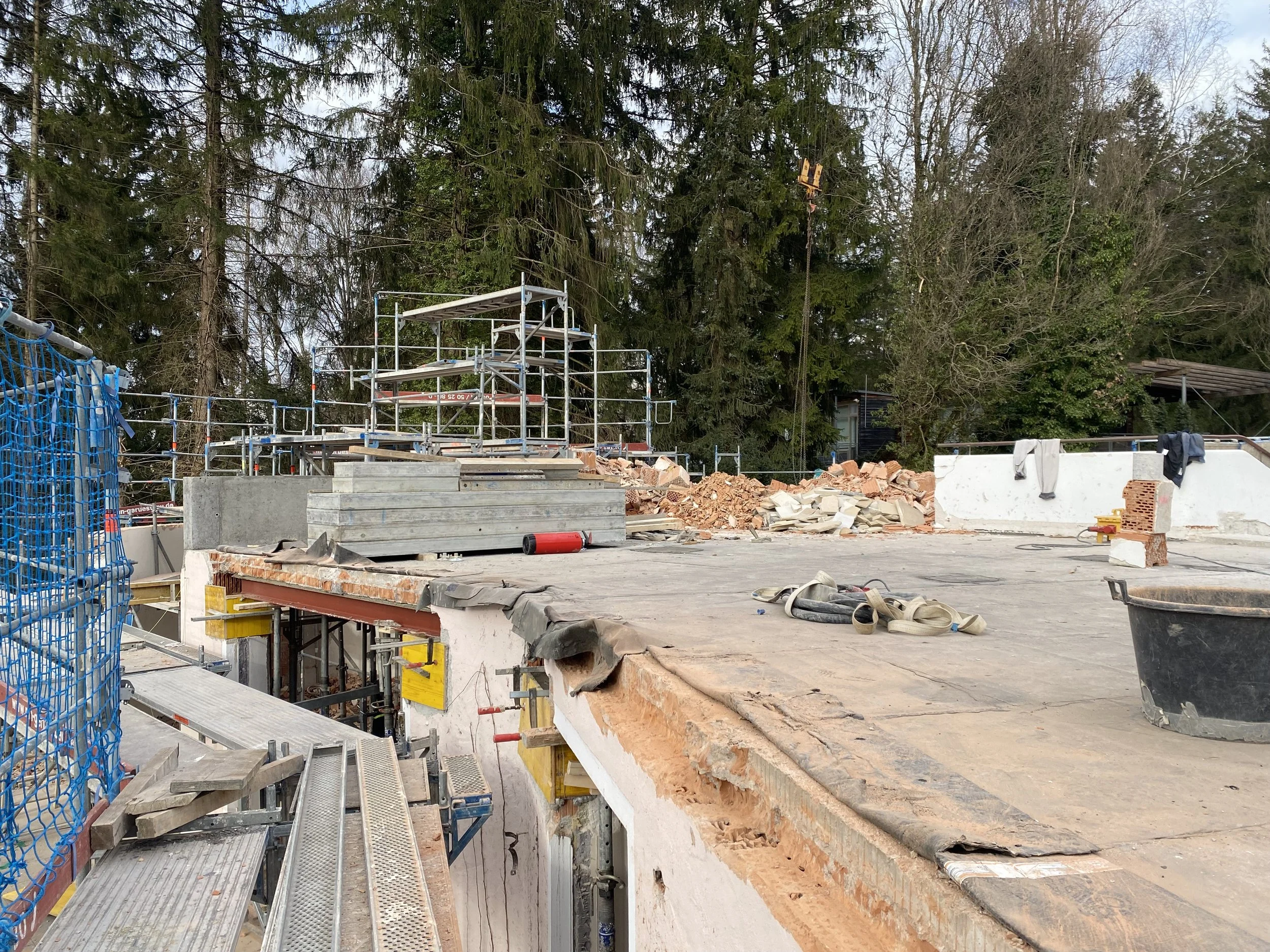 Baustelle mit Baugerüsten, Mauerreste, Werkzeuge und Baumstämme im Freien, umgeben von Bäumen.