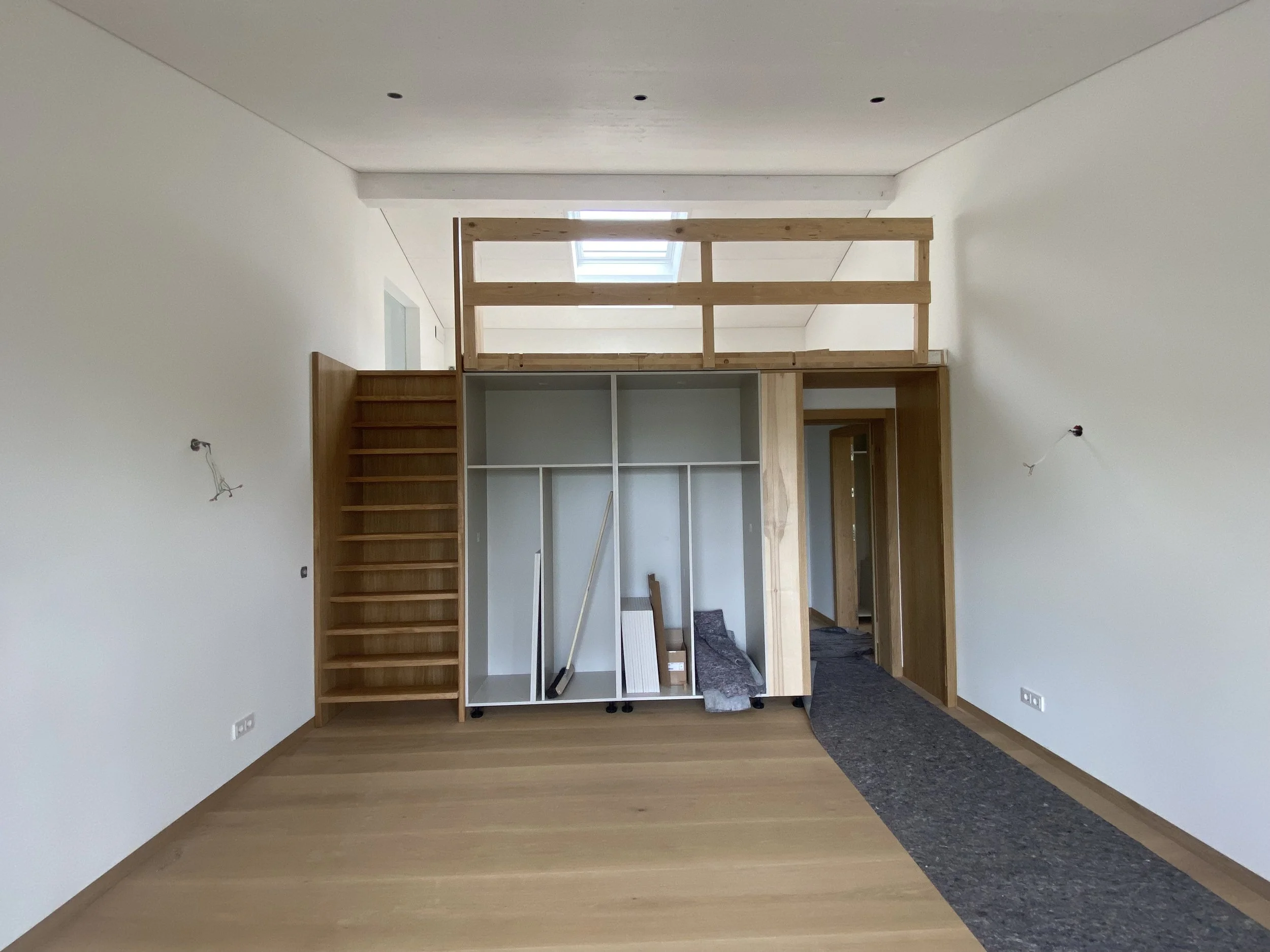 Innenraum eines fertiggestellten Raumes mit einer Holztreppe und einem Hochbett aus Holz, Holzfußboden, weiß gestrichenen Wänden und einem Fenster im Dach. Es gibt noch keine Möbel, nur Baumaterialien und Werkzeuge stehen im Raum.