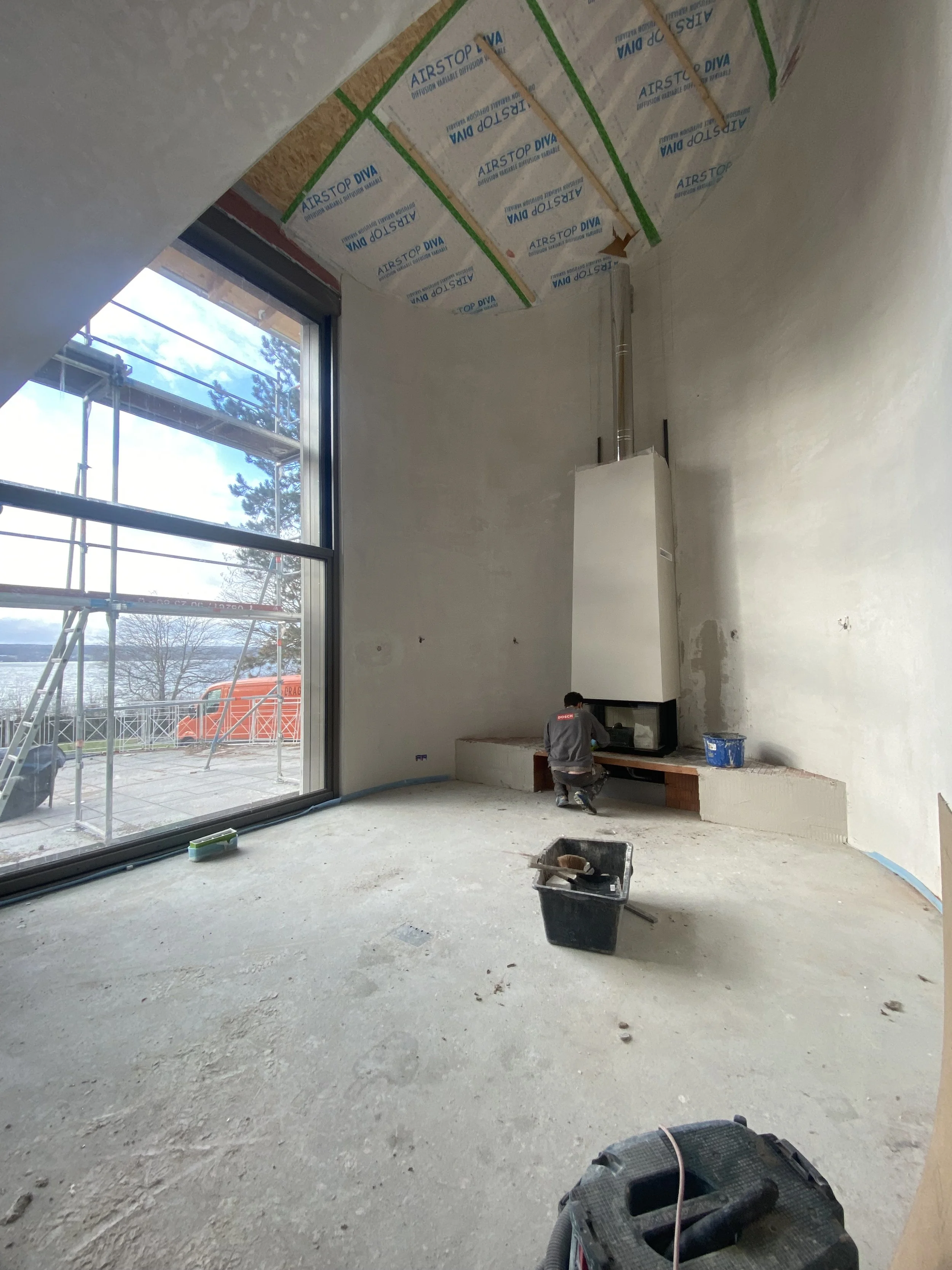 Raum im Bau mit großen Fenster, noch unbefliesteter Wand und einer Person, die an einem Kamin arbeitet. Es gibt Bauwerkzeug und Material im Raum.