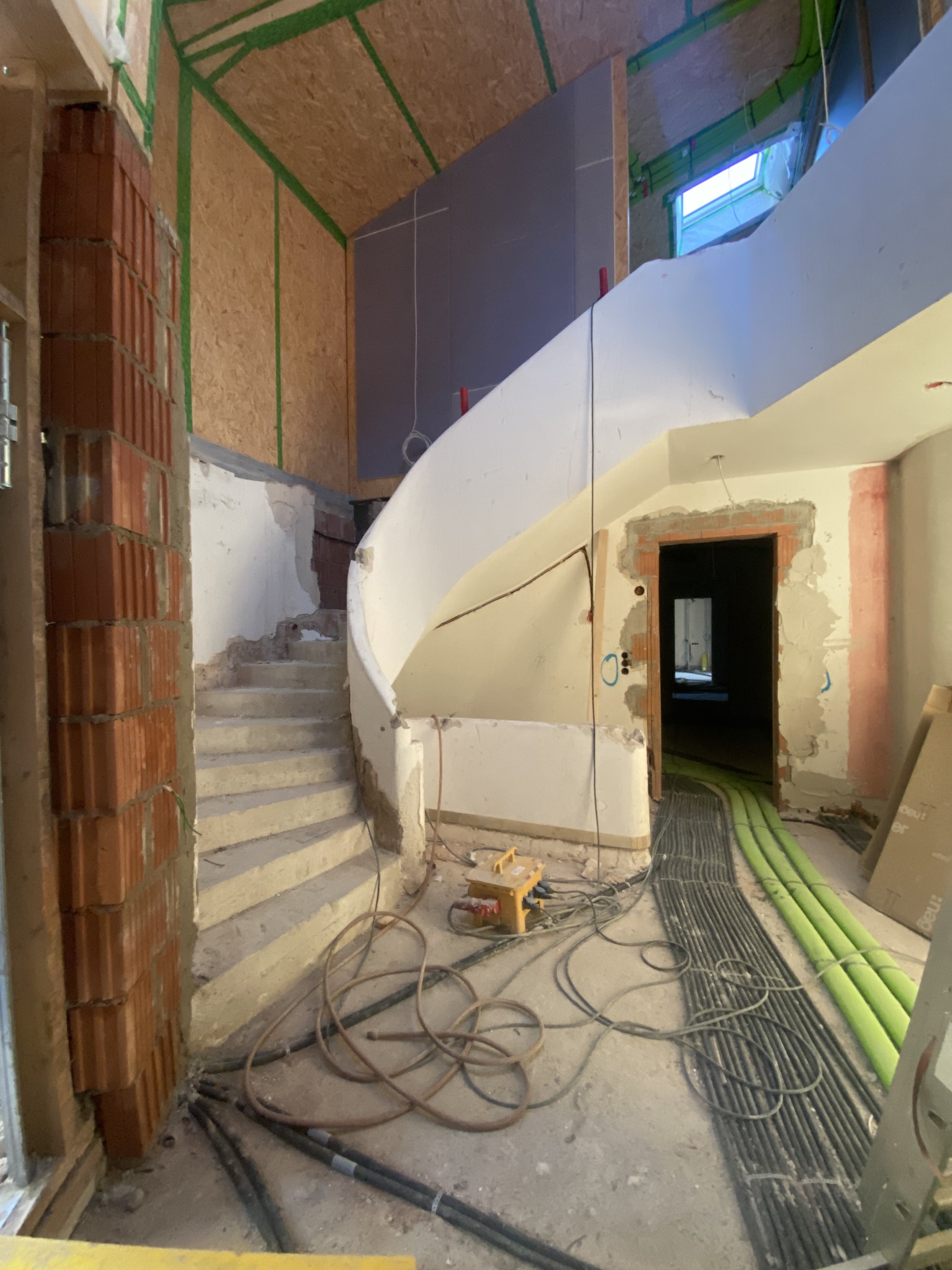 Innenansicht eines im Bau befindlichen Hauses mit rotem Ziegelstein, weißen Wänden und einer geschwungenen Treppe. Es gibt Bauwerkzeuge, Kabel und Rohre auf dem Boden. Eine Leiter ist im linken Bildrand sichtbar, und auf der oberen Ebene ist eine Wan