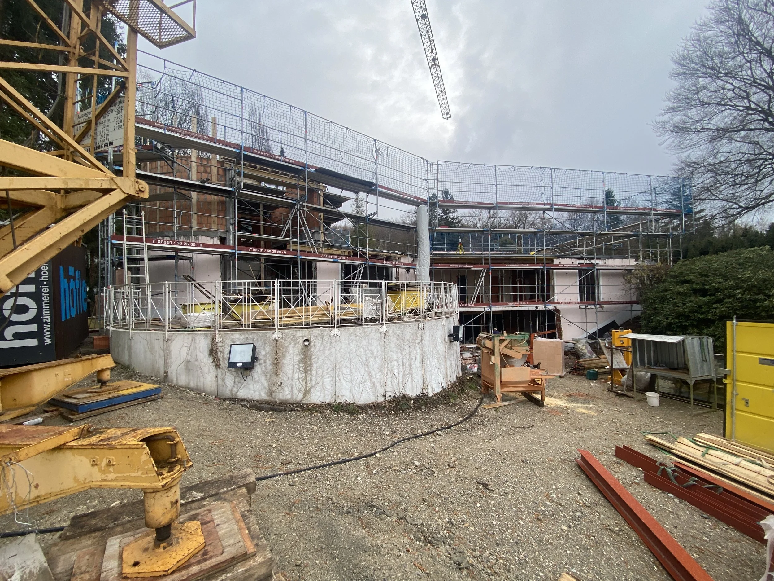 Baustelle mit Gerüst und Bauarbeiten an einem Gebäude, Baum im Hintergrund, Werkzeug und Baumaterial auf unbefestigtem Boden