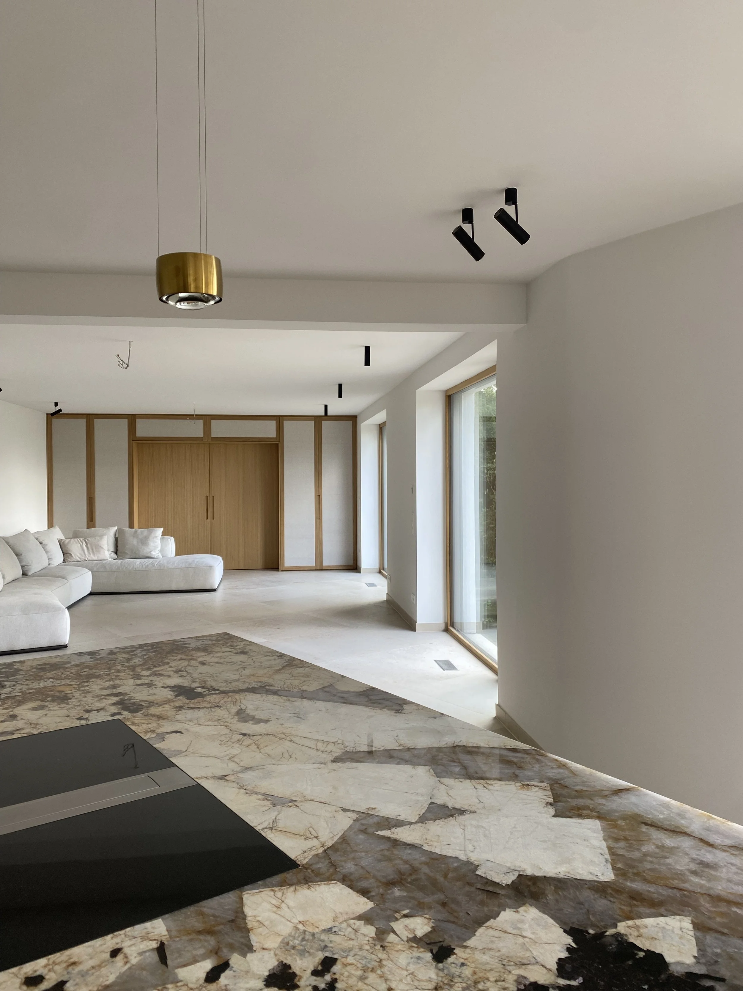 Moderner, heller Wohnraum mit Sofa, Holzschrank, Marmorarbeitsplatte, große Fenster, weiße Wände, verschiedene Deckenleuchten.