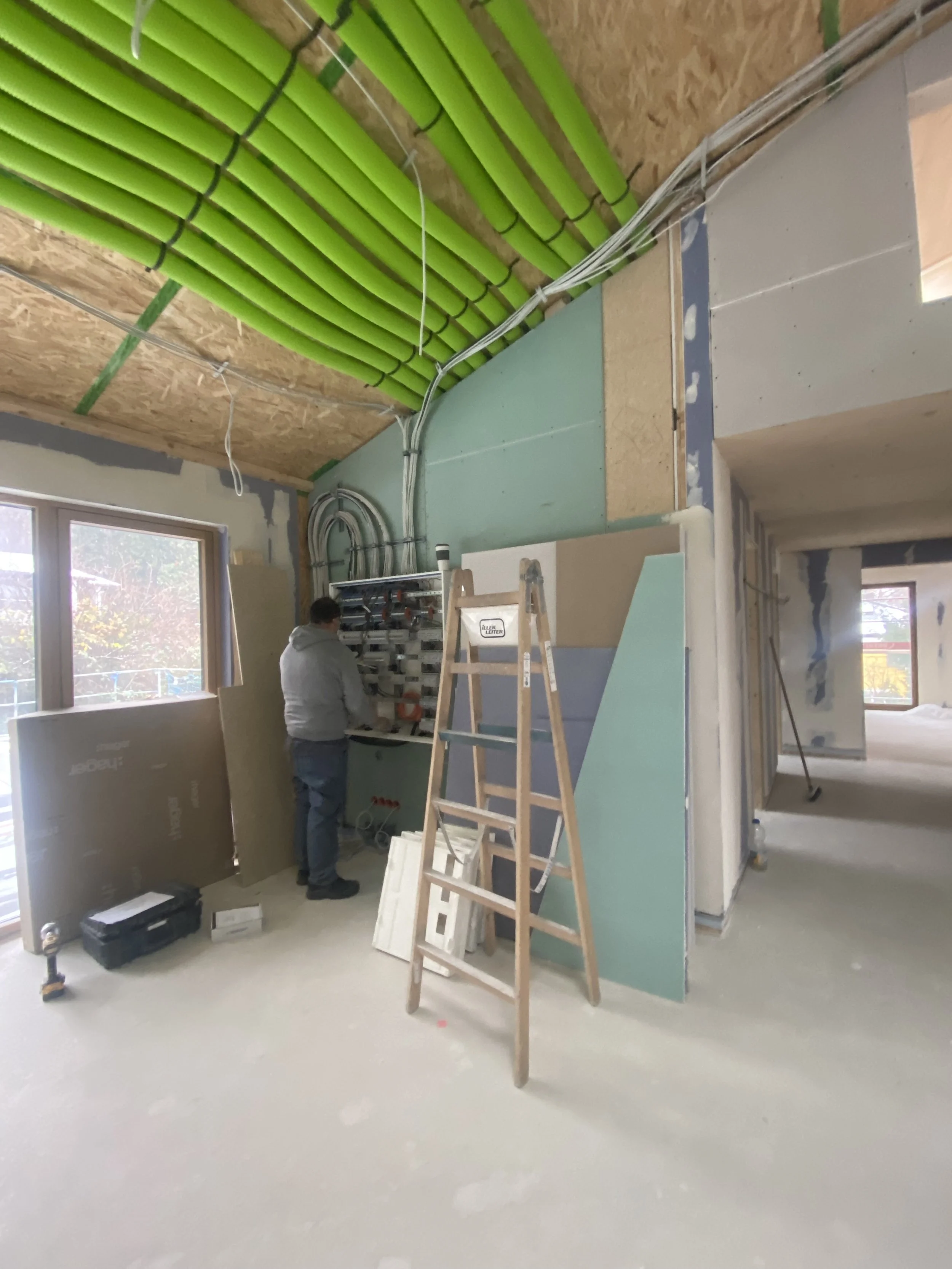 Innenaufnahme eines Gebäudes im Bau, mit einem Mann, der an einer Wand arbeitet. Im Raum sind Bauwerkzeuge, eine Leiter und Baumaterialien sichtbar, die Decke ist teilweise offen mit grünen Rohrleitungen.