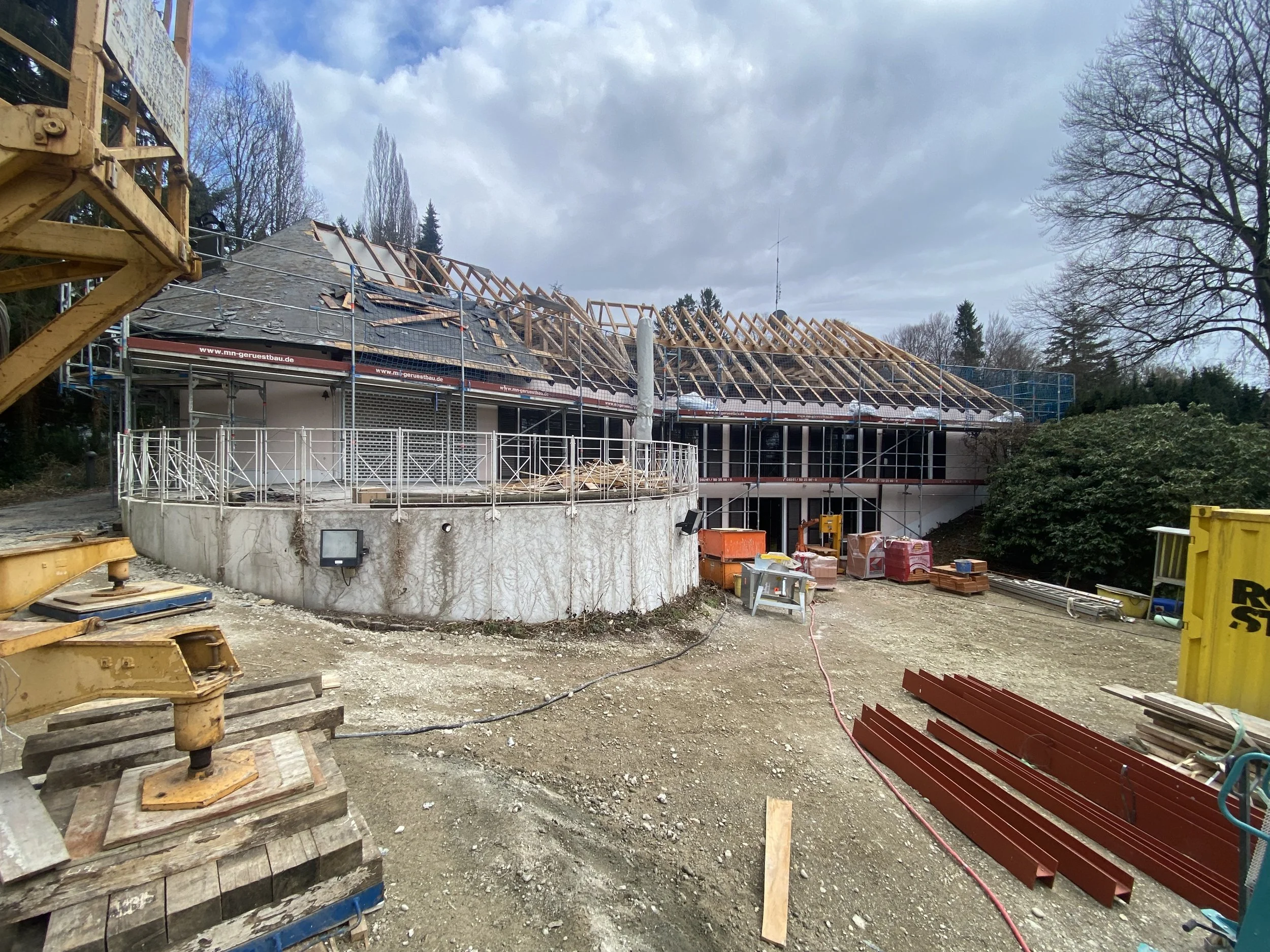Baumaßnahmen an einem Haus mit Holzdachkonstruktion, Gerüsten und Bauzubehör auf einem unbefestigten Gelände.