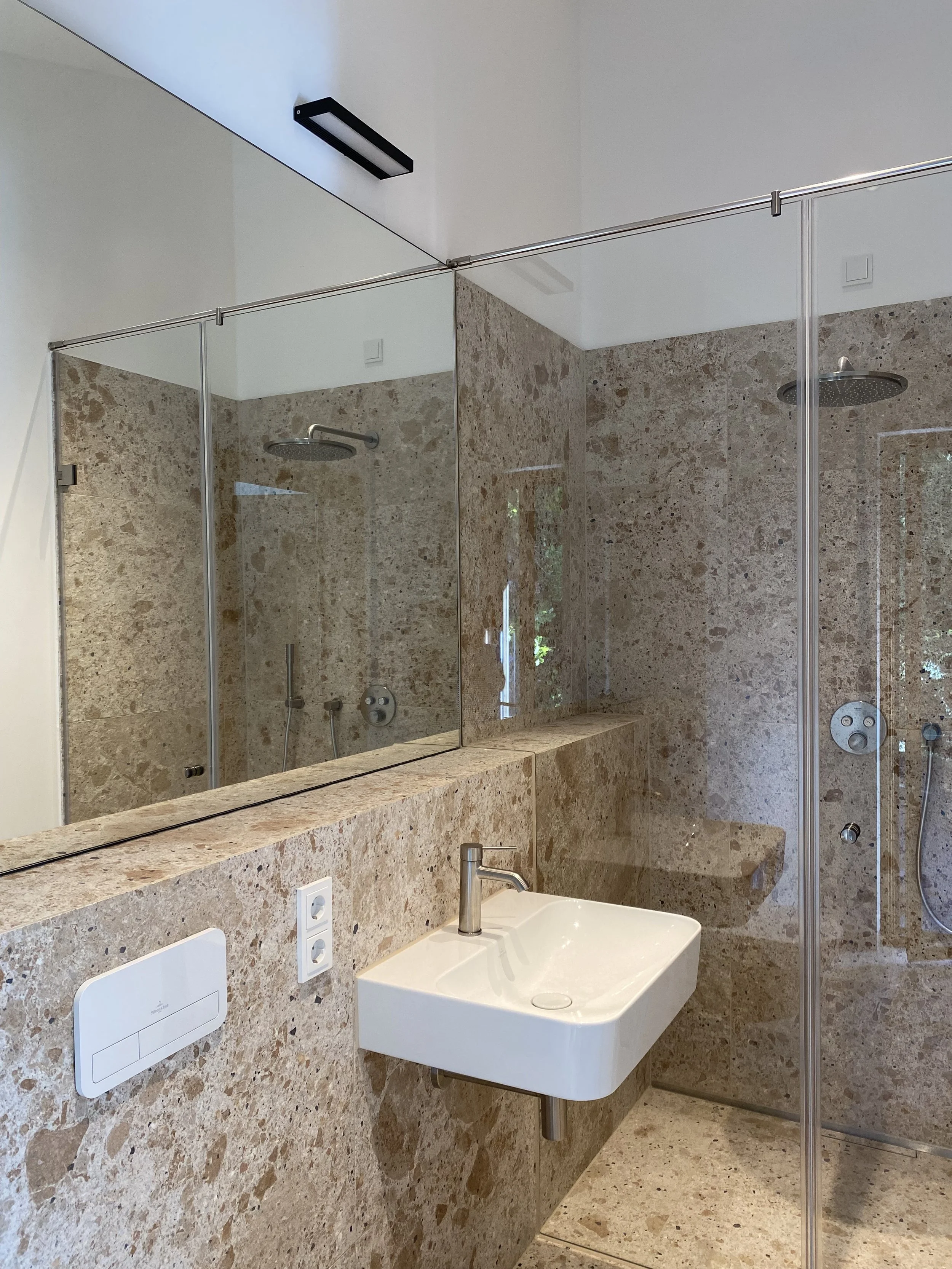 Ein Badezimmer mit einer Sandstein-Wand und -Boden, einer weißen Waschtisch und einem großen Spiegel, darüber eine schwarze Beleuchtung. Im Hintergrund sind eine Dusche mit Glaswand und eine kleine Sitzbank sichtbar.