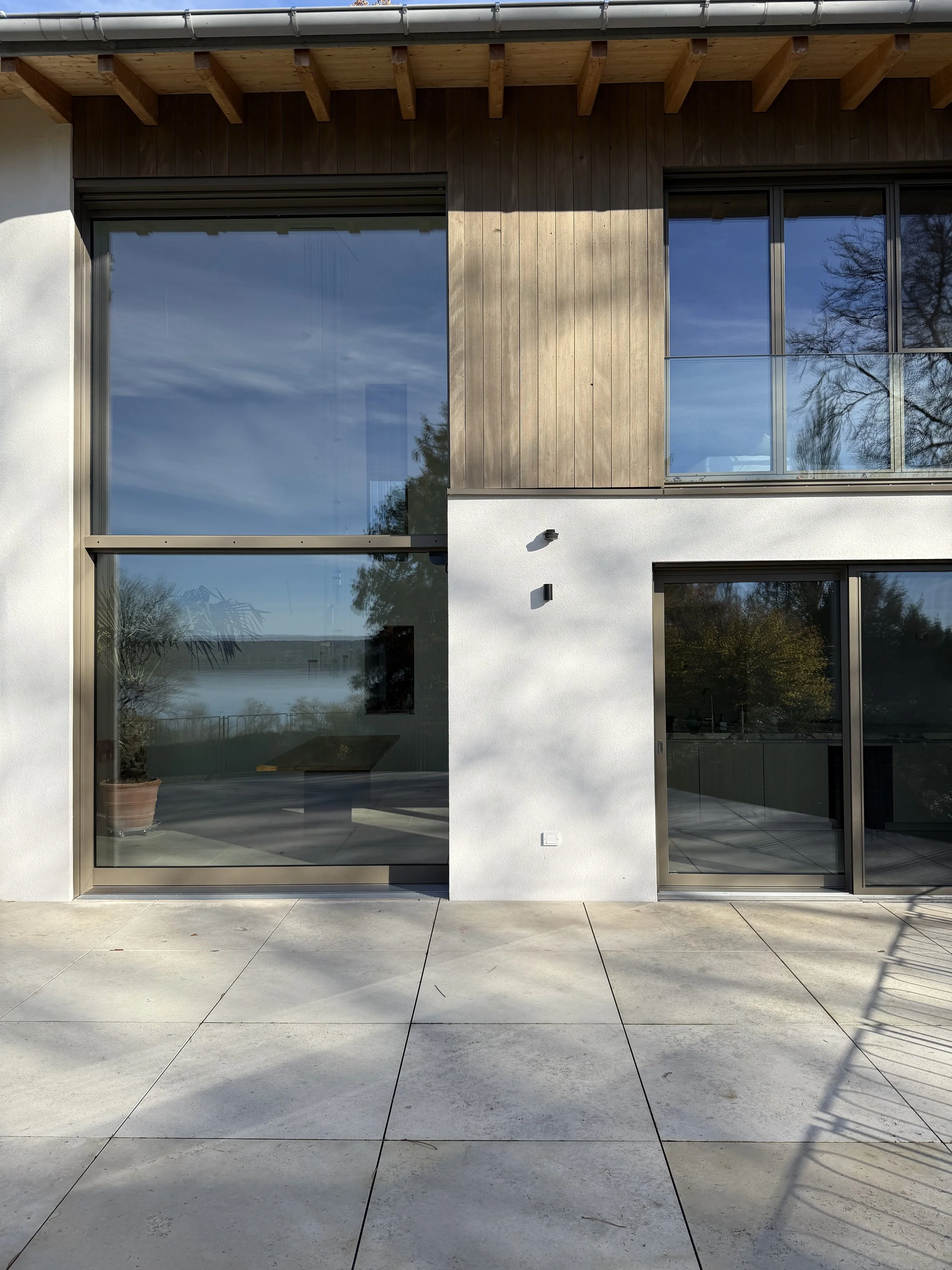 Ansicht einer modernen Gebäudefassade mit großen Glasfenstern, weißen und holzverkleideten Flächen, und einem Terrassenboden aus großformatigen, hellen Fliesen.