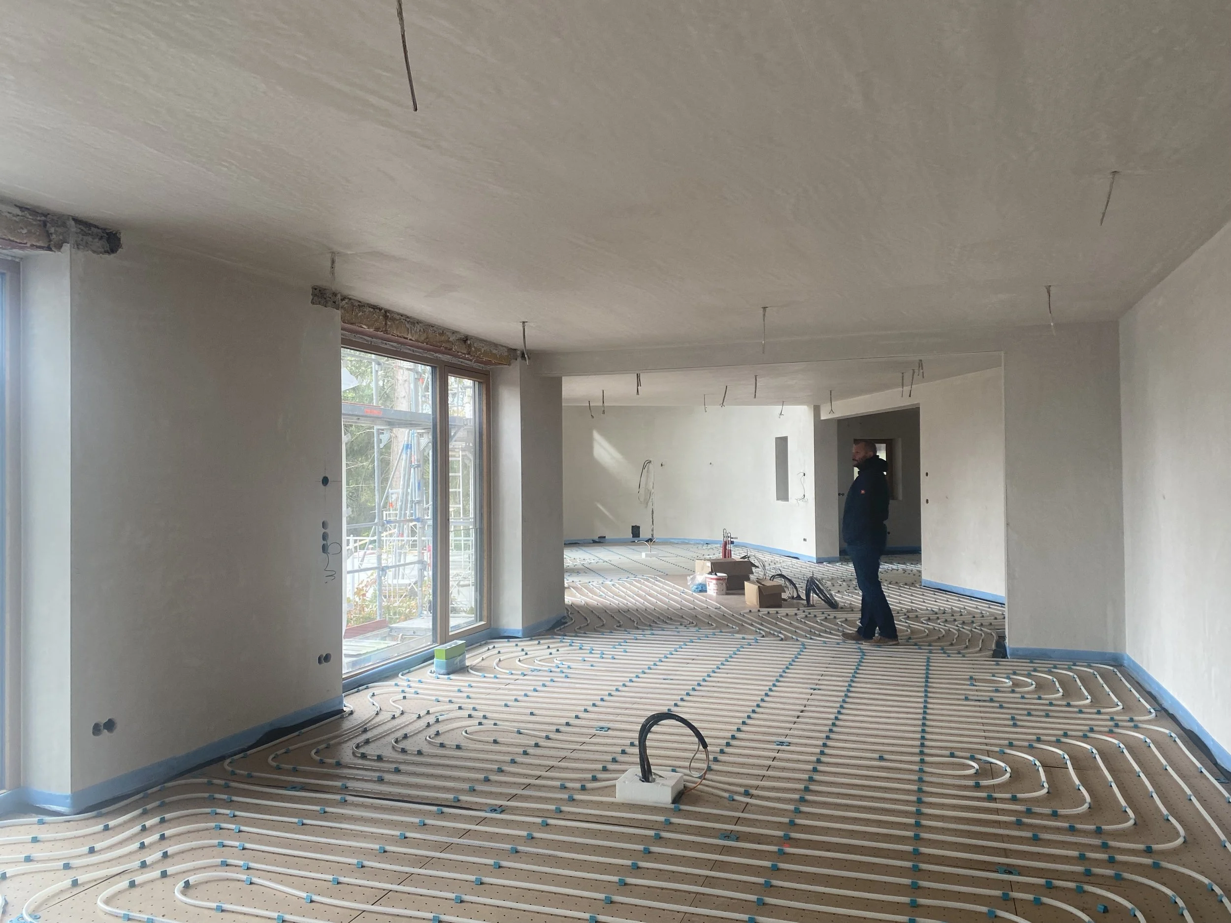 Innenaufnahme eines unfertigen Wohnzimmers im Bau, mit Heizungsrohren auf dem Boden, einer Person, die die Baustelle inspiziert, und großen Fenstern, die nach draußen führen.