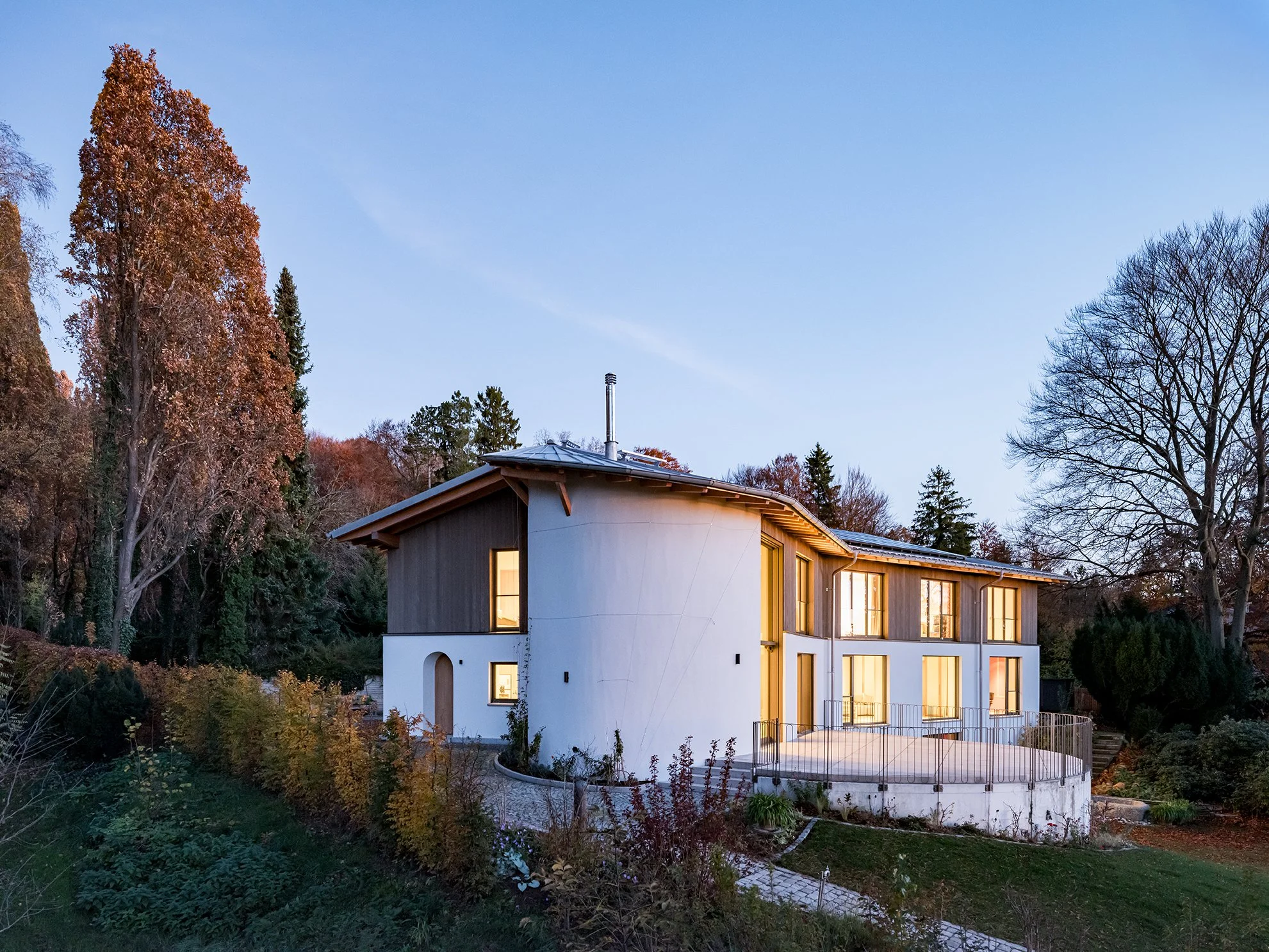 Modernes Haus mit Holz- und Weißfassade, große Fenster, auf einem Hang mit Garten bei Sonnenuntergang, umgeben von Bäumen.
