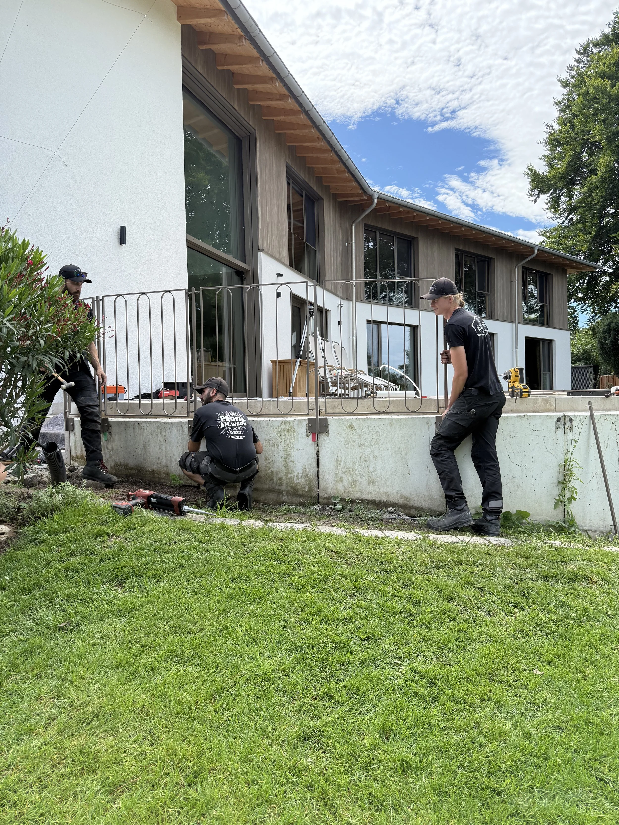 Drei Männer arbeiten an der betonierten Umrandung eines Hauses im Garten. Sie tragen schwarze Arbeitskleidung und benutzen Werkzeuge. Das Haus hat große Fenster und eine moderne Fassade.