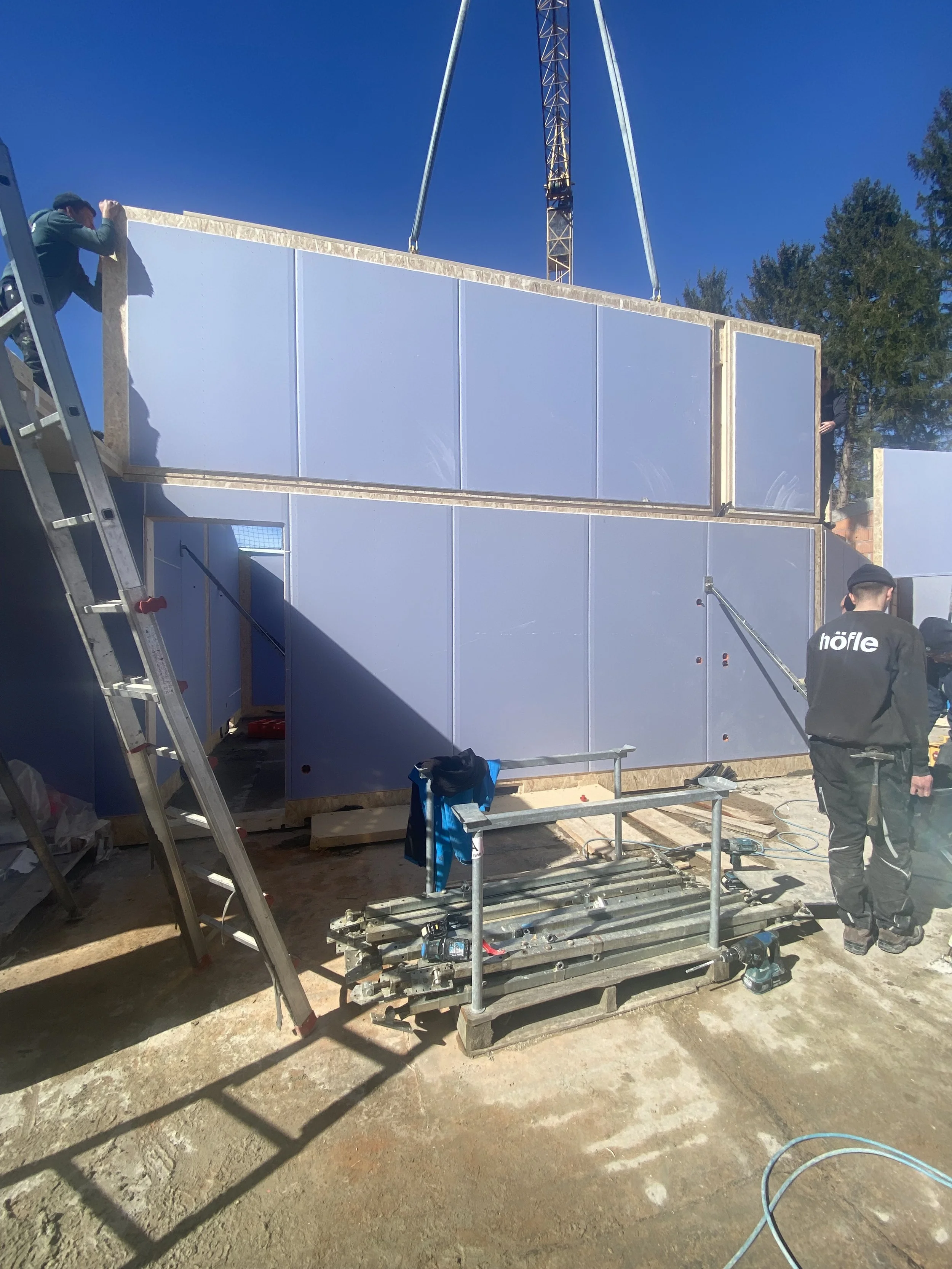 Bauarbeiter errichten eine Wand mit blauen Wandpaneelen auf einer Baustelle im Freien, bei sonnigem Himmel.