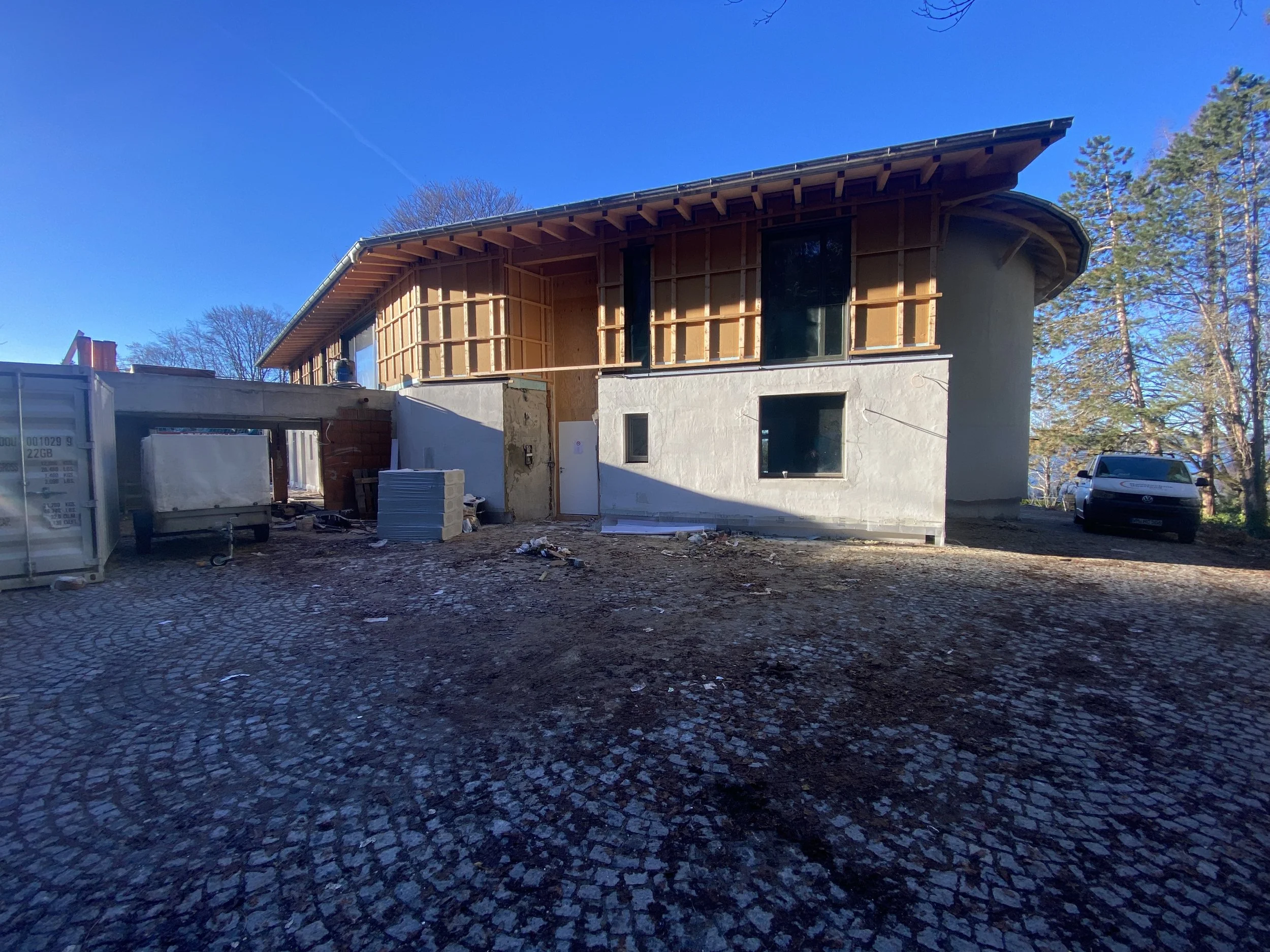 Ein Haus im Bau mit unvollständiger Fassade, Baumaterialien im Vordergrund, ein Auto auf der rechten Seite und eine Baumgruppe im Hintergrund, alles unter klarem blauen Himmel.