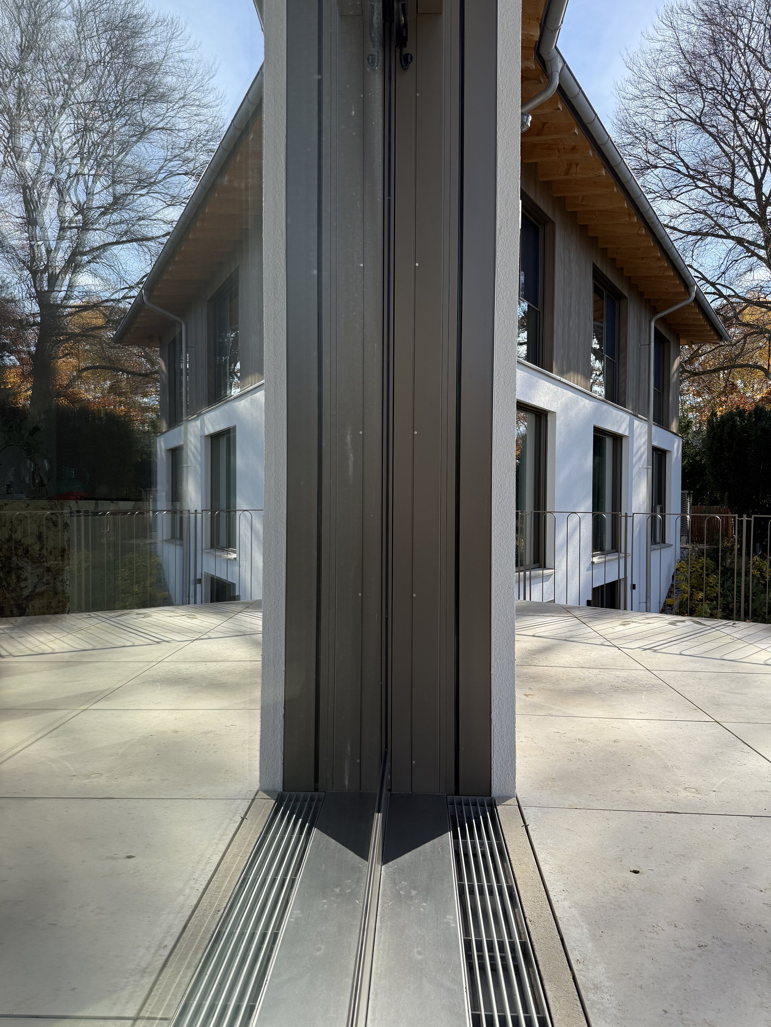 Ansicht eines modernen Gebäudes mit großen Fenstern, Dachüberstand aus Holz, seitlichem Geländer und gläserner Tür, mit Blick auf den Garten und größere Bäume im Hintergrund.