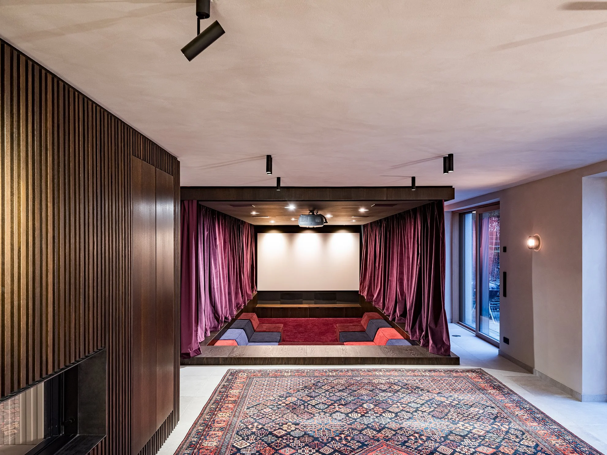 Ein moderater Heimkinoraum mit einer weiße Leinwand, roten Vorhängen und einer niedrigen Sitzreihe mit Kissen auf einem erhöhten Podest, neben großen Glastüren, die nach draußen führen.