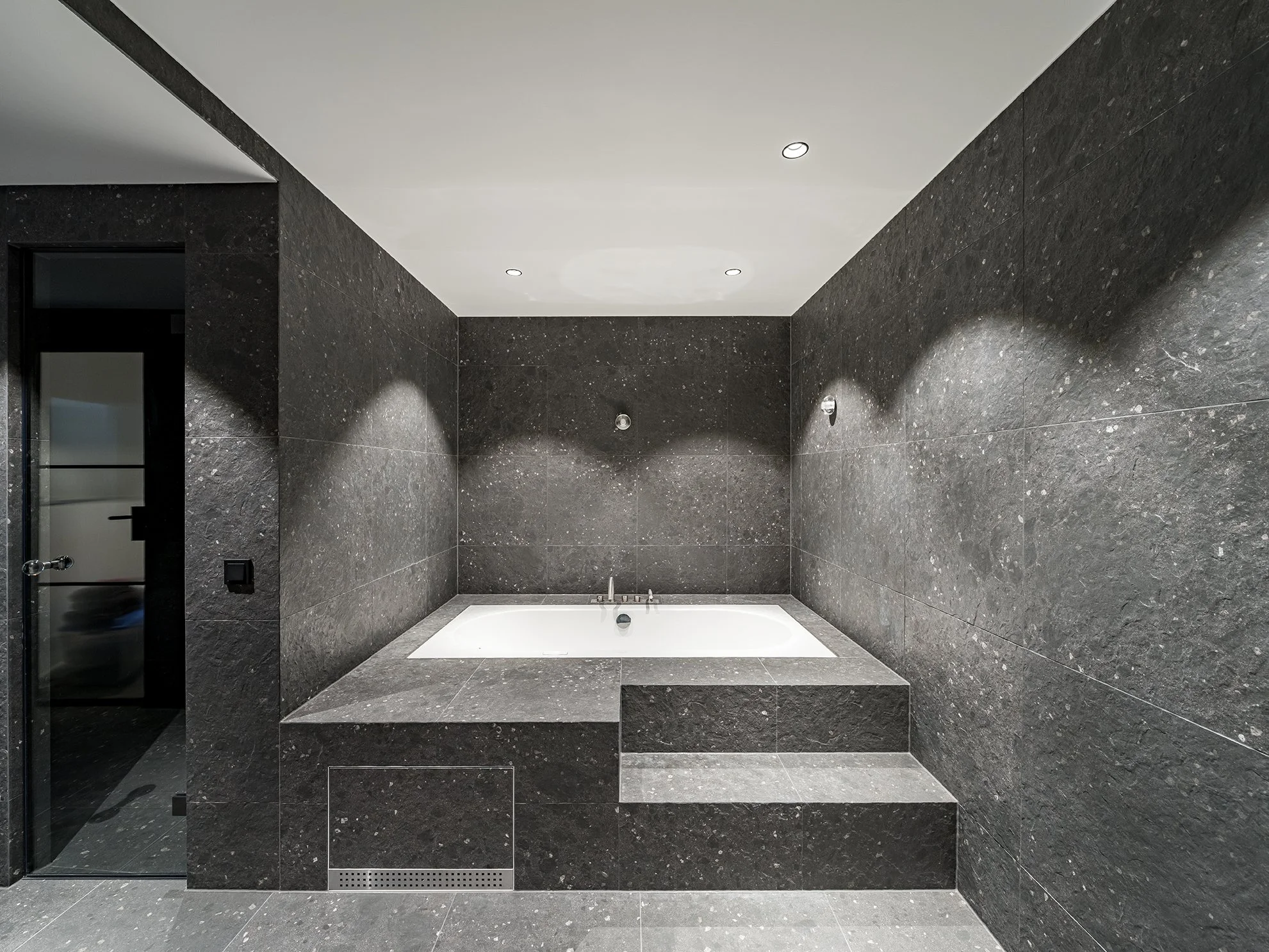 Moderner Wellness-Badezimmer mit schwarzer Granitverkleidung, Badewanne auf Plattform und Dusche im linken Vordergrund.