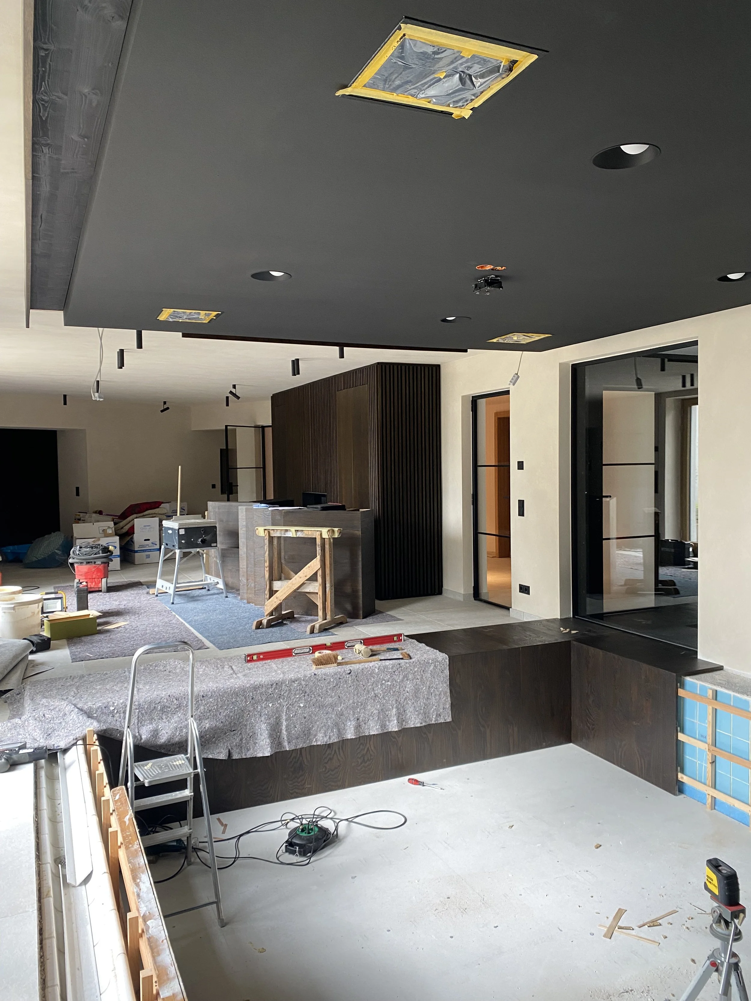 Innenaufnahme eines Hauses im Bau, mit dunklem Deckensystem, teilweise fertiggestellten Wänden, Baustellenwerkzeugen und Baumaterialien.