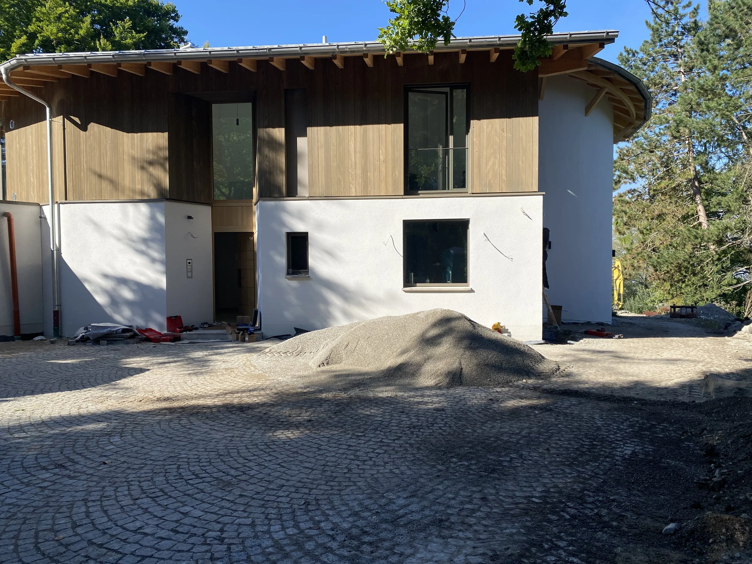 Ein modern gebautes Haus unter Bauarbeiten, mit einem gepflasterten Vorplatz und Baustoffen im Vordergrund, umgeben von Bäumen.