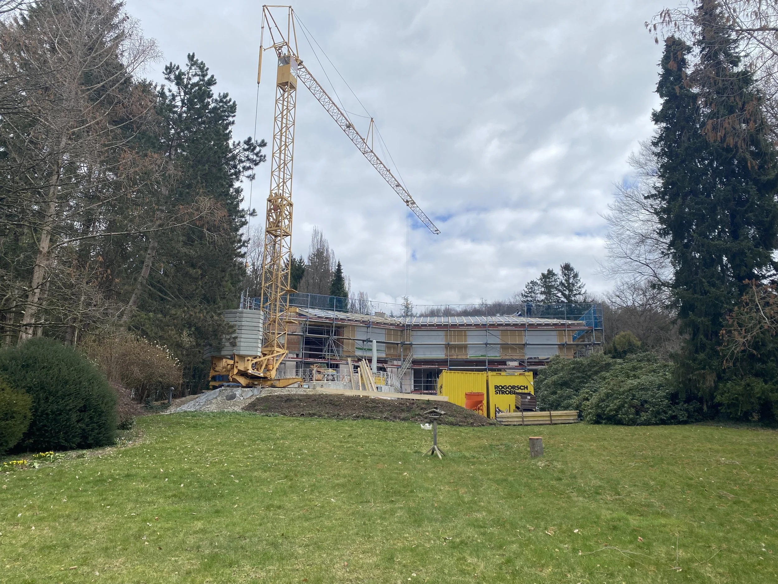 Ein Baukran auf einer Baustelle, im Hintergrund ein Gebäude im Bau, umgeben von Bäumen und einem grünen Rasen im Vordergrund.