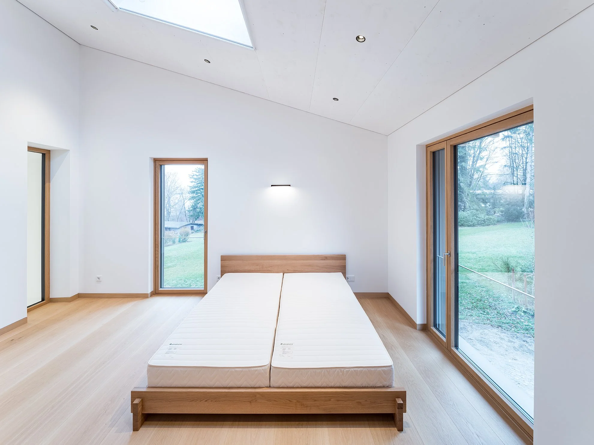 Schlafzimmer mit weißem Wand, Holzfußboden, zwei große Fenster und ein Doppelbett ohne Bettwäsche, Blick nach draußen auf grüne Wiese und Bäume.
