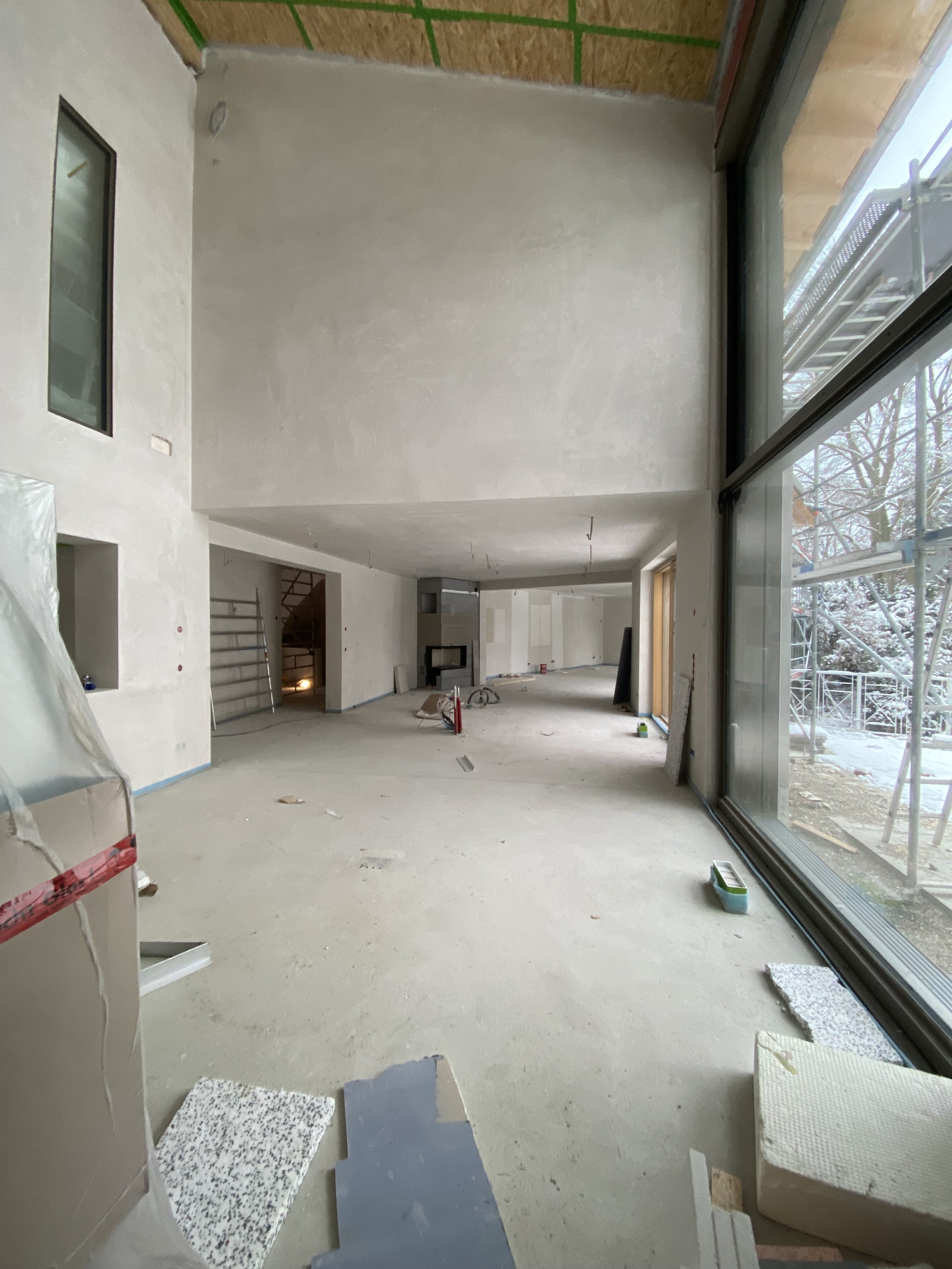 Innenraum eines Hauses im Bau, ungefertig, große Fensterfront auf der rechten Seite, offen gestaltet mit Terrasse außerhalb, Baustellenschritte sichtbar.