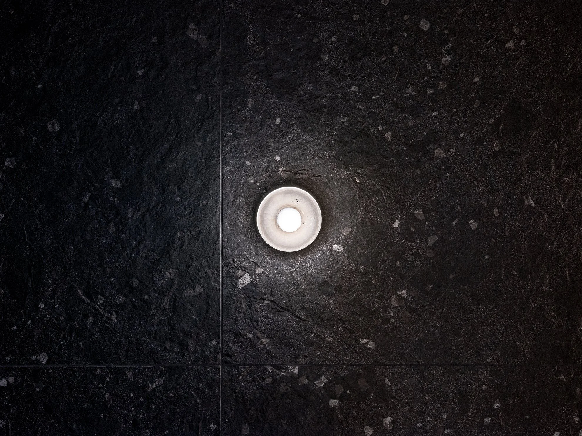 Ein runder grauer Gegenstand auf einer schwarzen, strukturierten Wand, mit einer hellen Mitte, die nach oben zeigt.